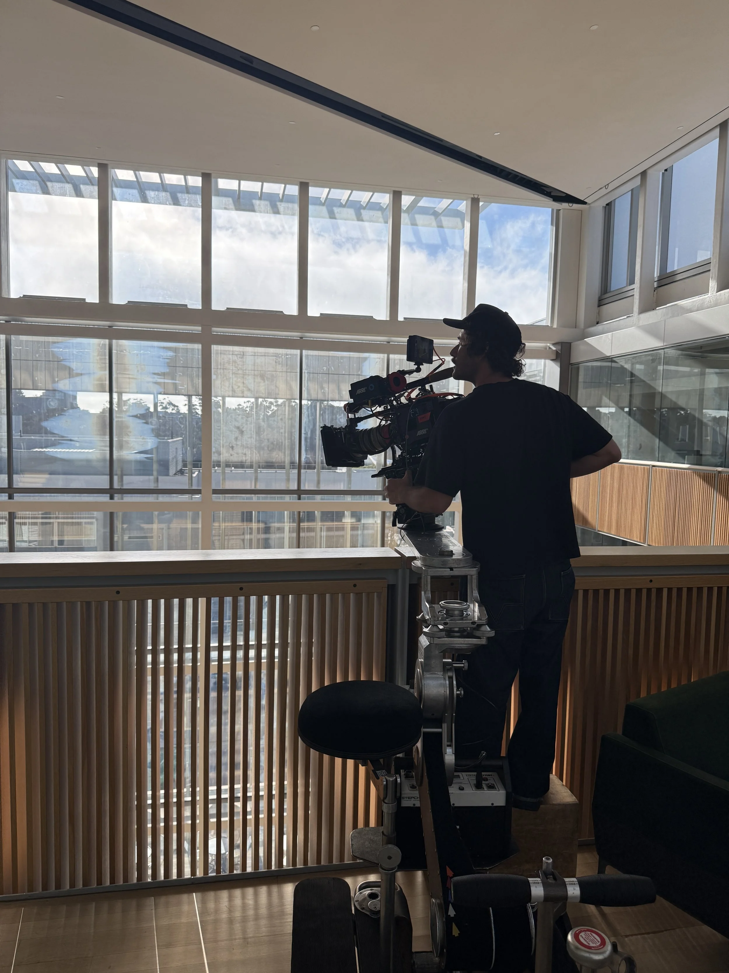 A person filming with a camera mounted on a steady-cam rig in an interior space with large windows and wooden paneling.