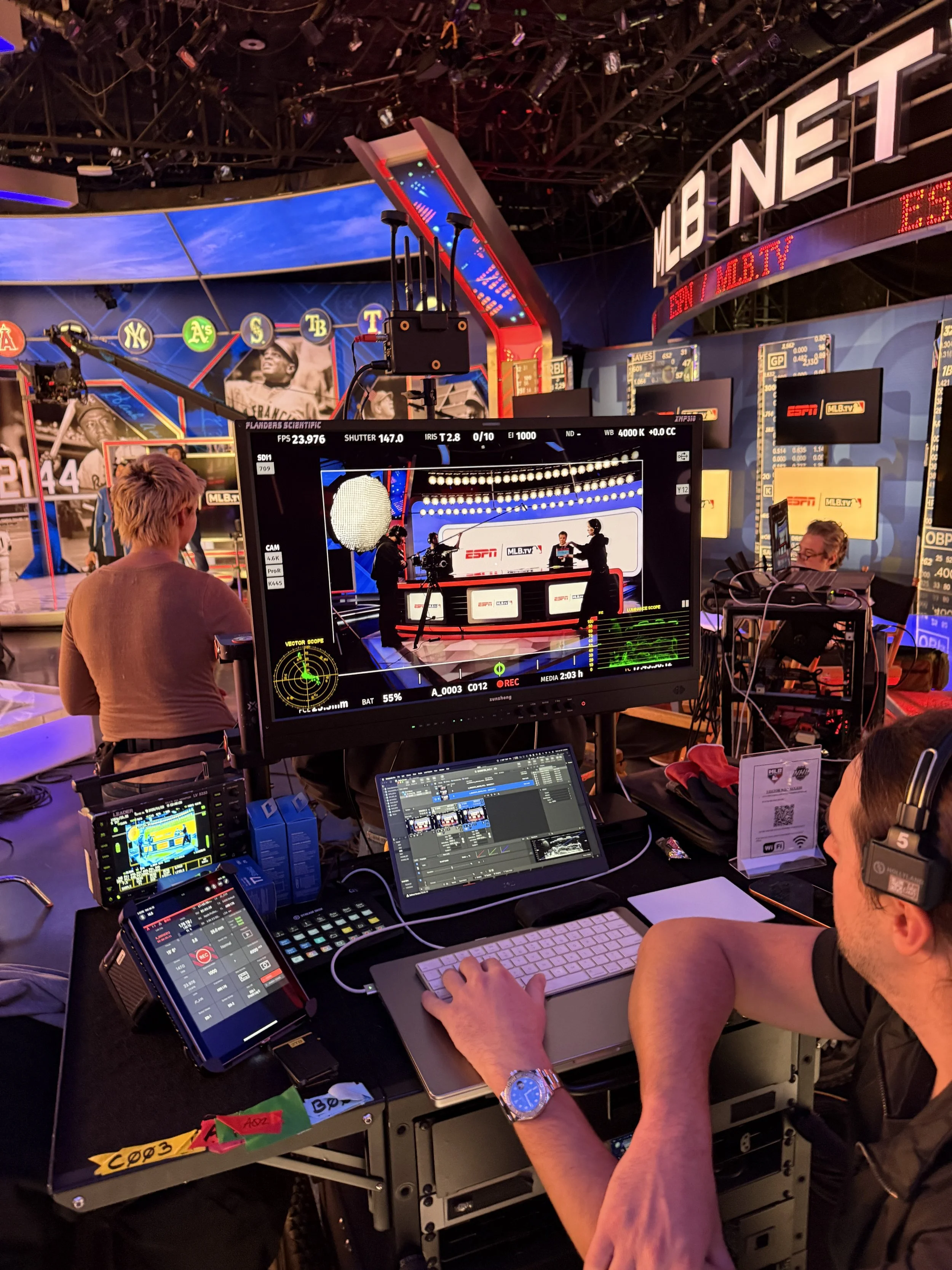 Behind-the-scenes view of a television studio with multiple monitors, cameras, and production equipment. Crew members are working on the set that features a large 'MLB NETWORK' sign and sports images on the wall.
