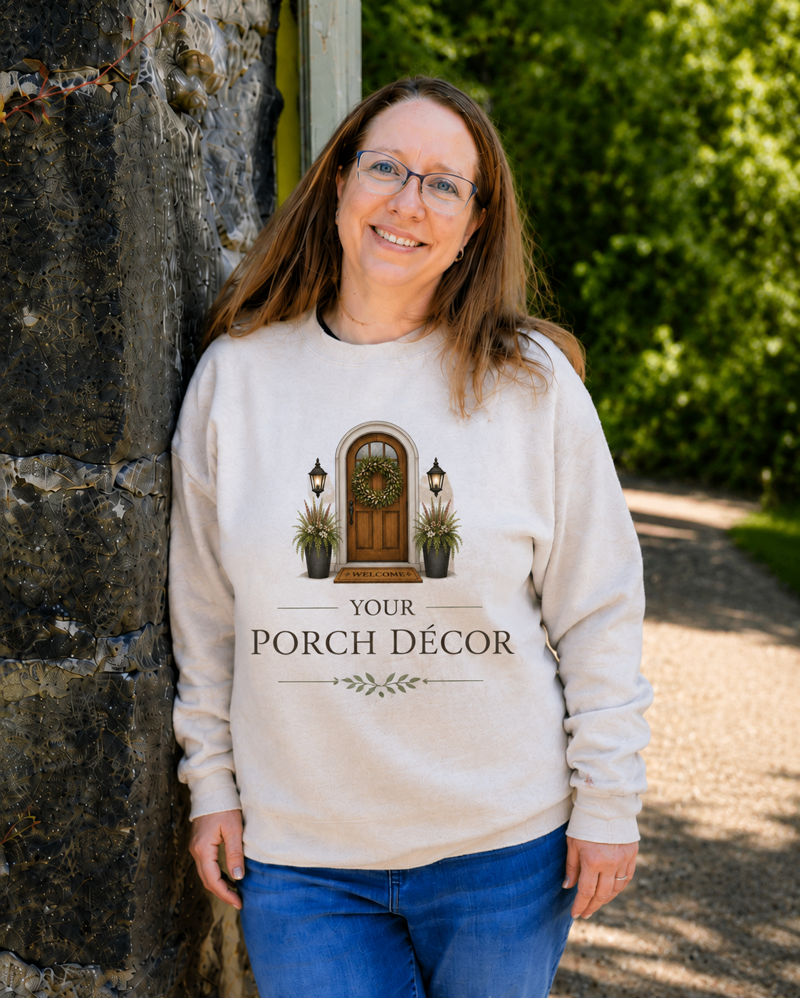 Woman smiling outdoors in front of a wall, wearing a white sweatshirt that has an image of a porch with a door and plants, and the text 'Your Porch Décor'.