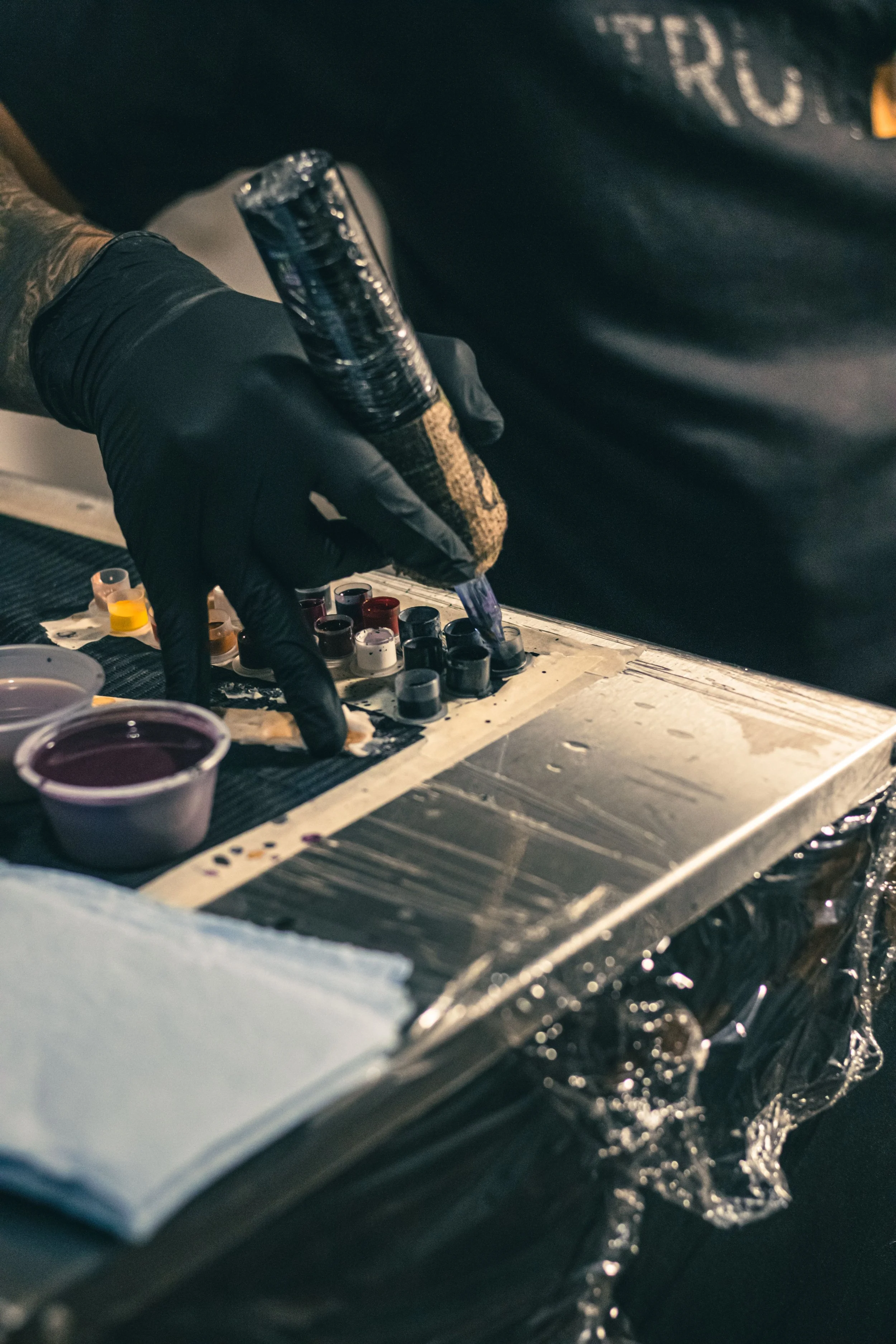 A person wearing black gloves holding a tattoo machine, tattooing on a surface. There are small containers of ink nearby, and the workspace appears to be covered with protective paper or foil.