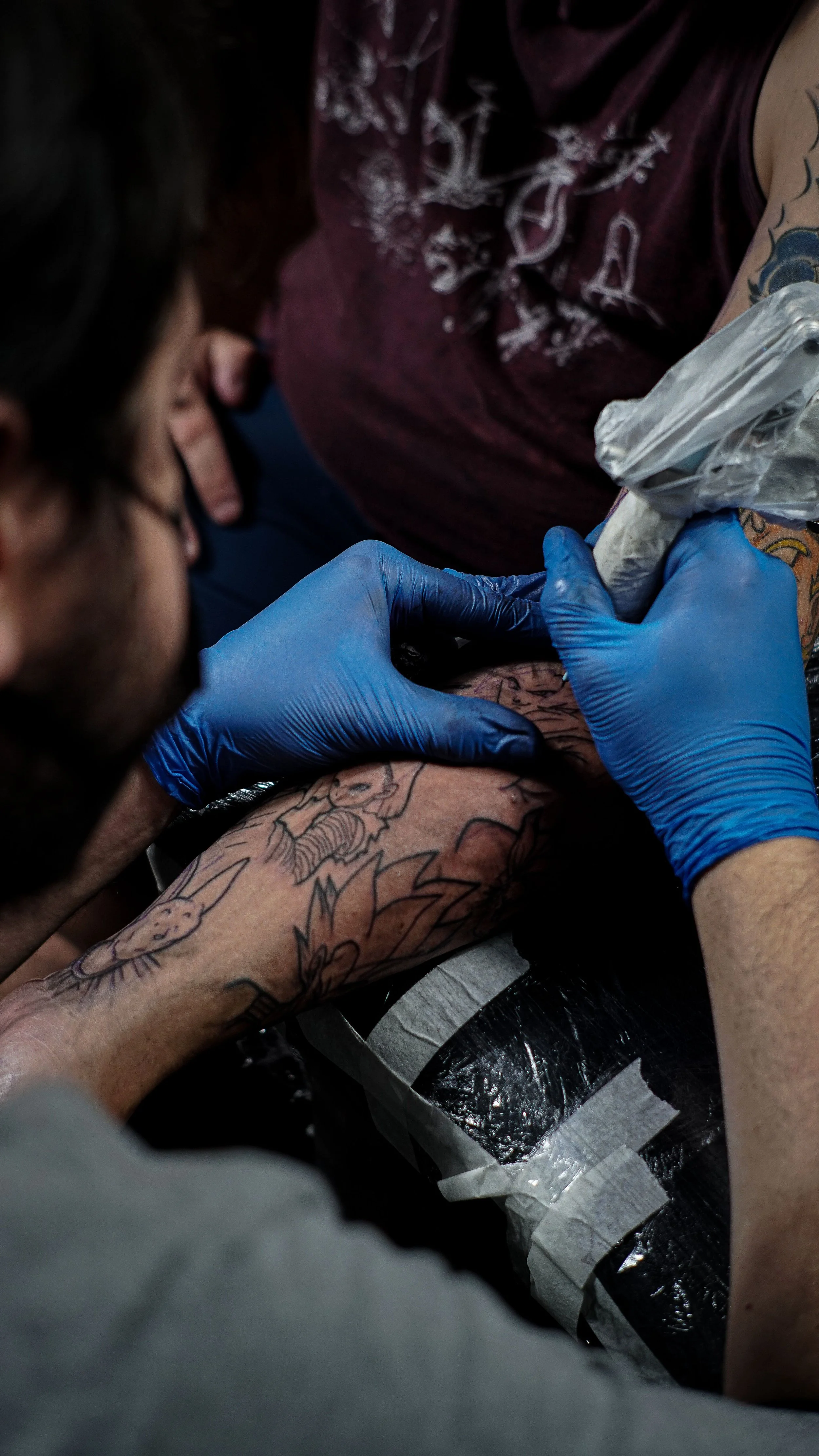 A person with tattoos getting a tattoo on their arm in a tattoo shop. The tattoo artist is using a tattoo machine, wearing blue gloves, and working on a detailed design.