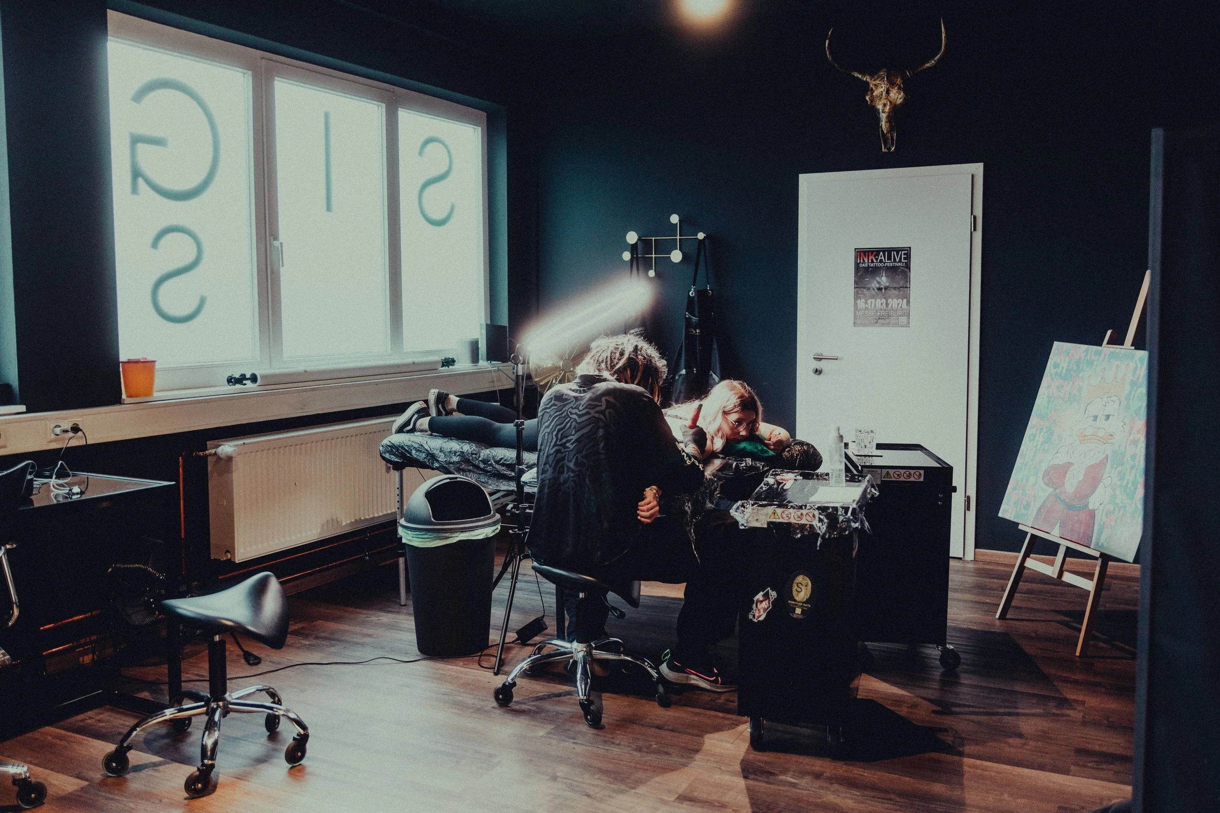 A room with dark blue walls and wooden flooring, decorated with a bull skull mounted on the wall above a white door, and a large window with frosted glass displaying the word 'SICS'. There are three people sitting around a table, two of them are lying on chairs and one sitting, surrounded by various objects including a canvas on an easel, and a tall standing lamp. A painting leaning against the wall depicts a clown.