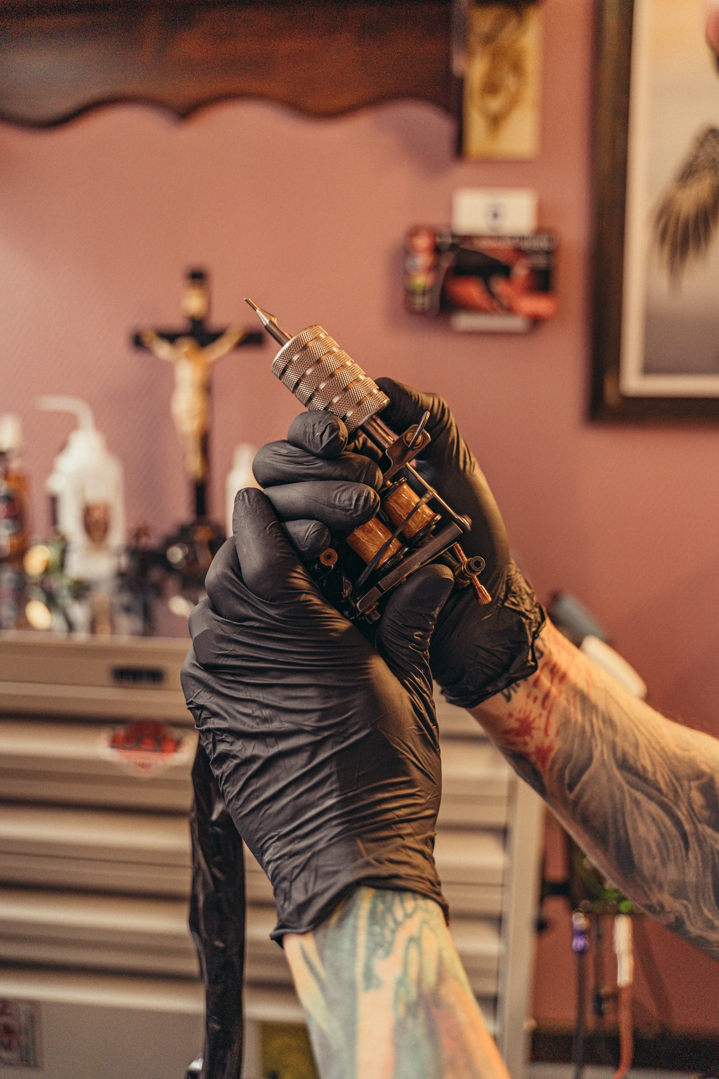 A person wearing black gloves holding a tattoo machine in a tattoo studio, with tattoo artwork on their arm and a background including religious statues and wall art.