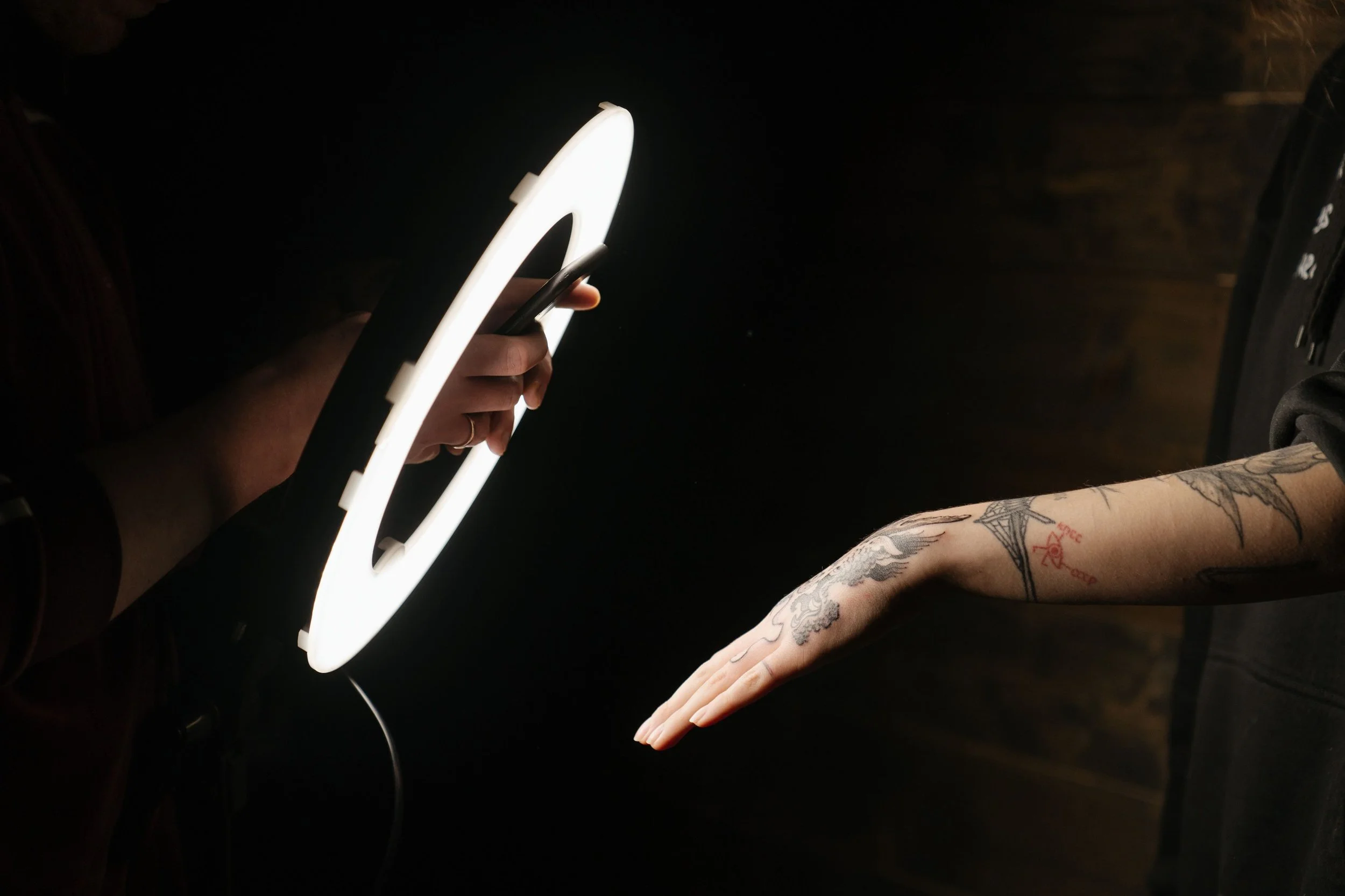 A person with tattoos on their forearm is resting their hand on a surface while another person holds a glowing neon sign shaped like a ring or a circle in front of their hand at night or in a dark setting.