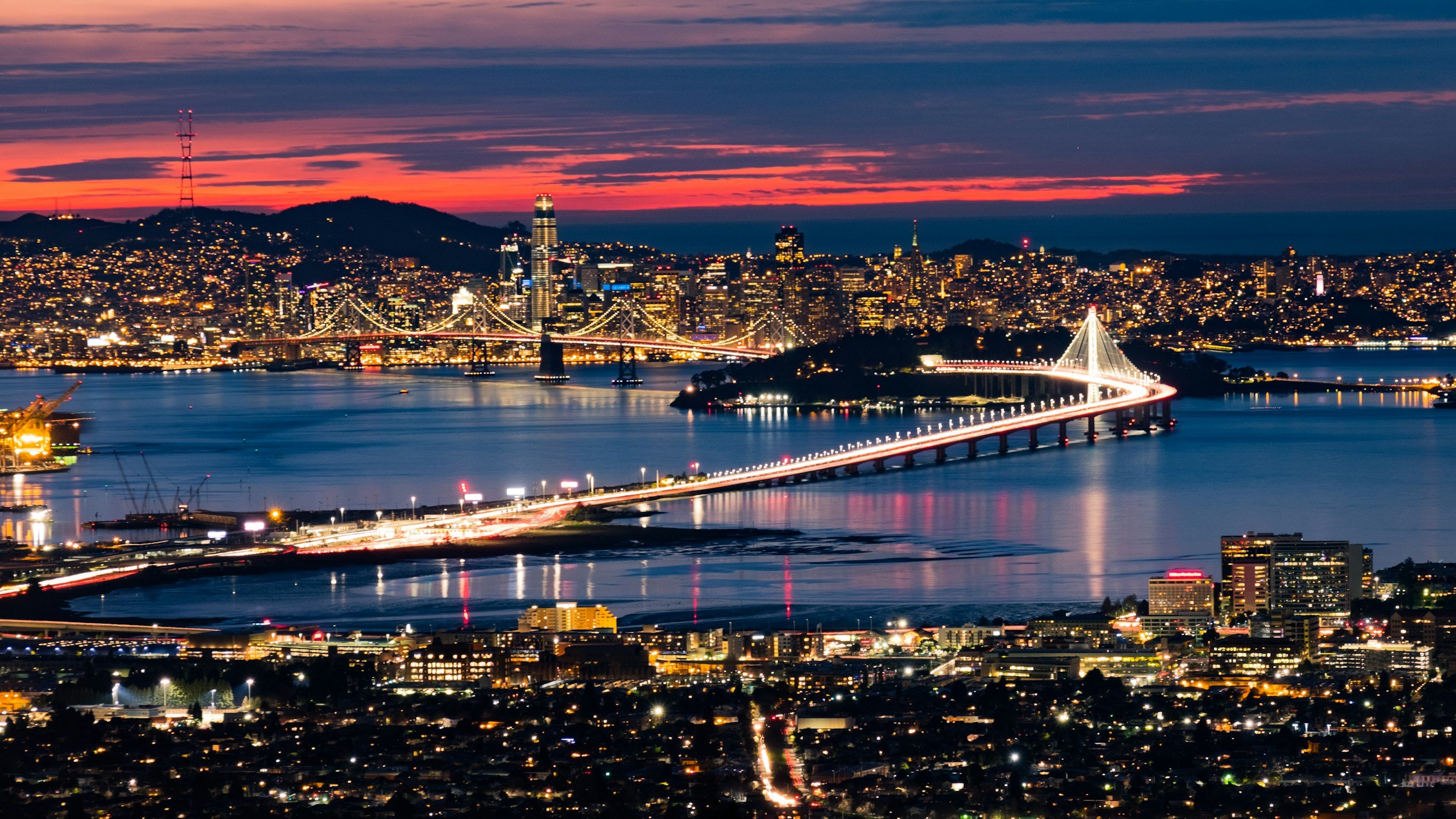 Night cityscape of San Francisco with illuminated bridges and skyscrapers, including the Transamerica Pyramid, over the bay with sunset sky.