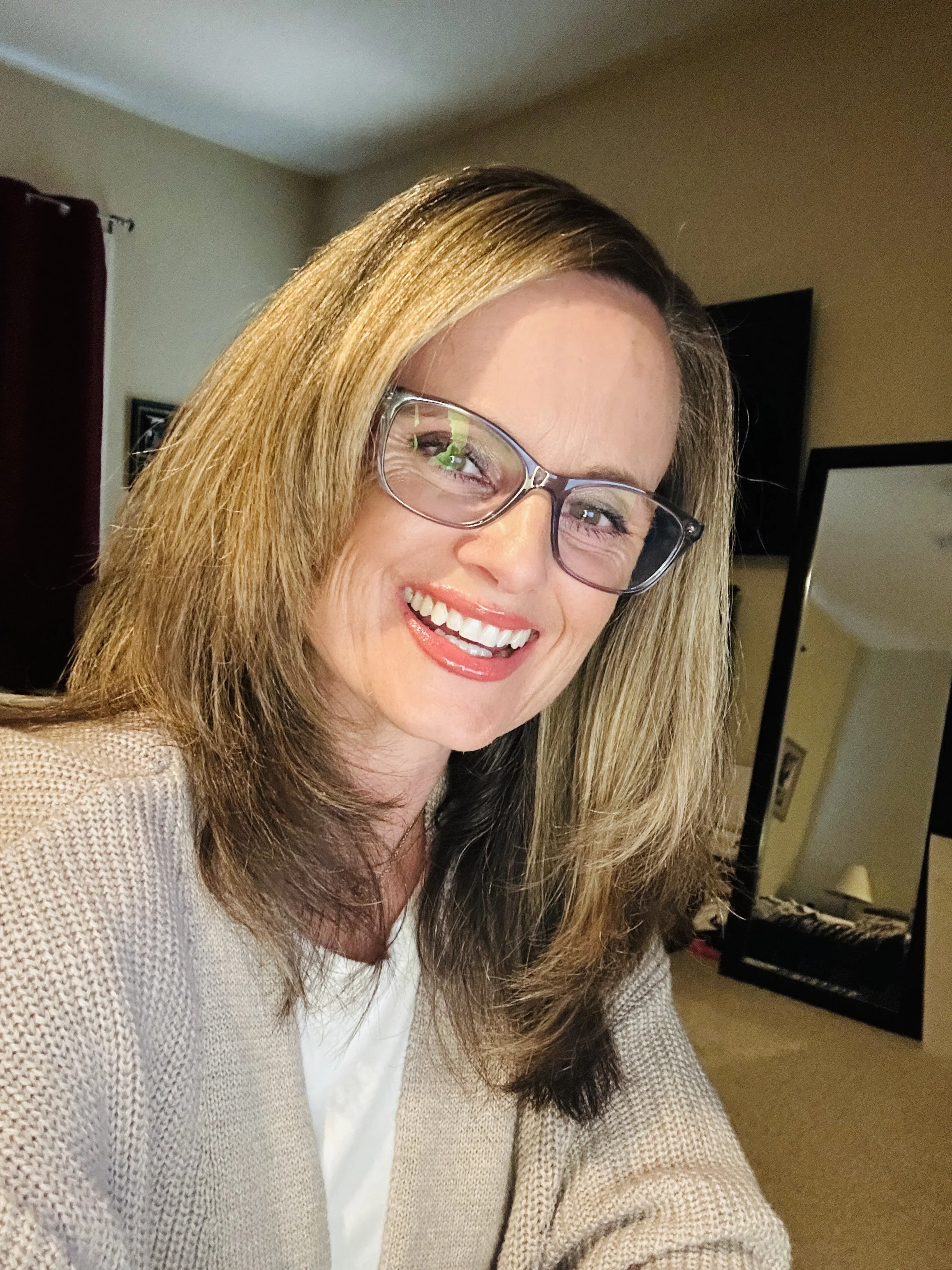 A smiling woman with light brown hair wearing glasses, a white shirt, and a beige cardigan in a living room.