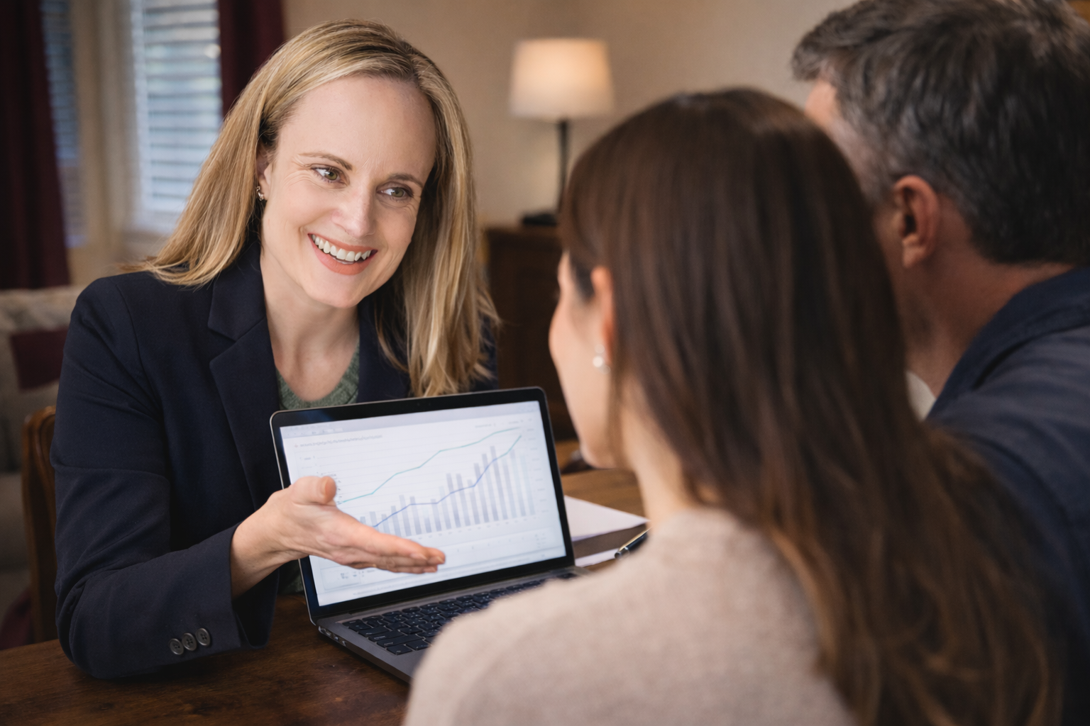 A woman in a dark blazer showing two people a laptop screen with graphs during a business meeting in a home office.