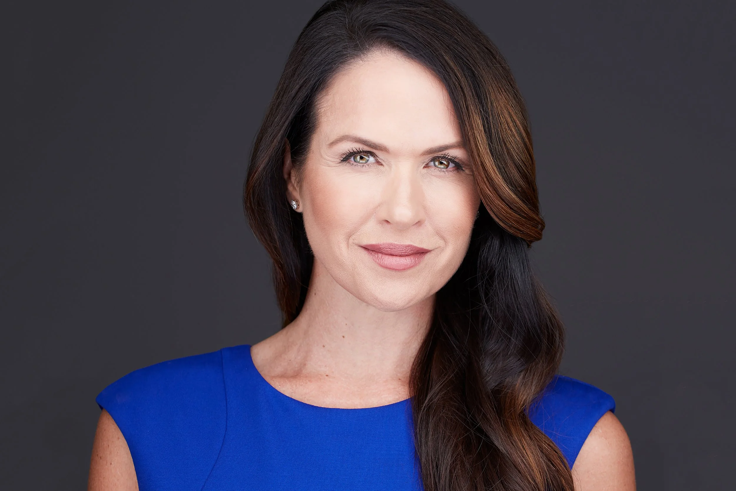 Portrait of a woman with long, dark hair, wearing a blue sleeveless dress, smiling slightly, photographed against a dark grey background.