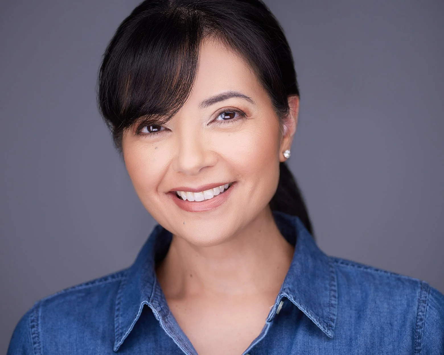 A woman with dark hair styled in a side part and a smile, wearing a blue denim shirt against a plain gray background.