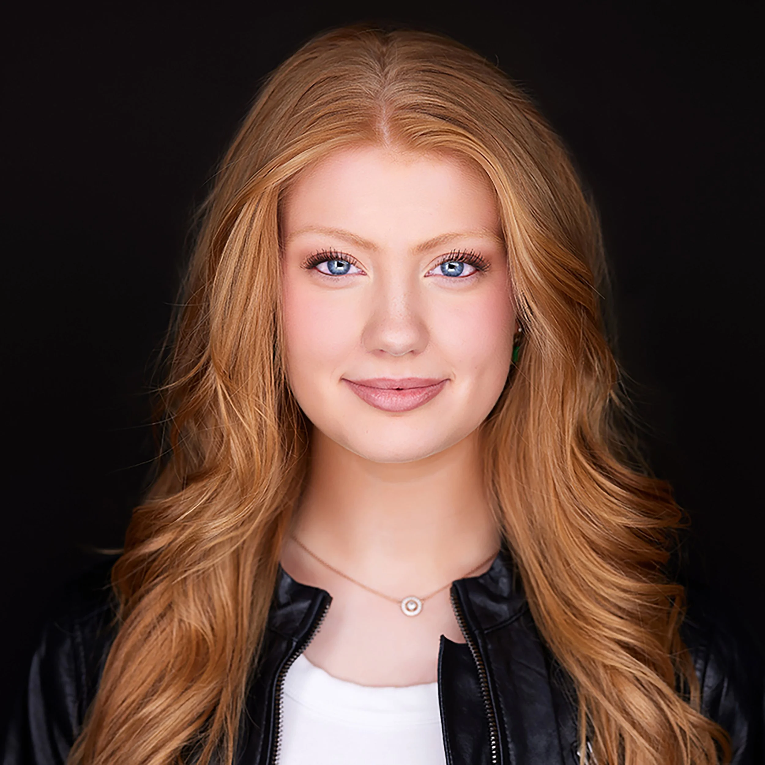 Close-up of a woman with red hair and blue eyes, wearing a black leather jacket, white top, and a necklace with a circular pendant, against a black background.