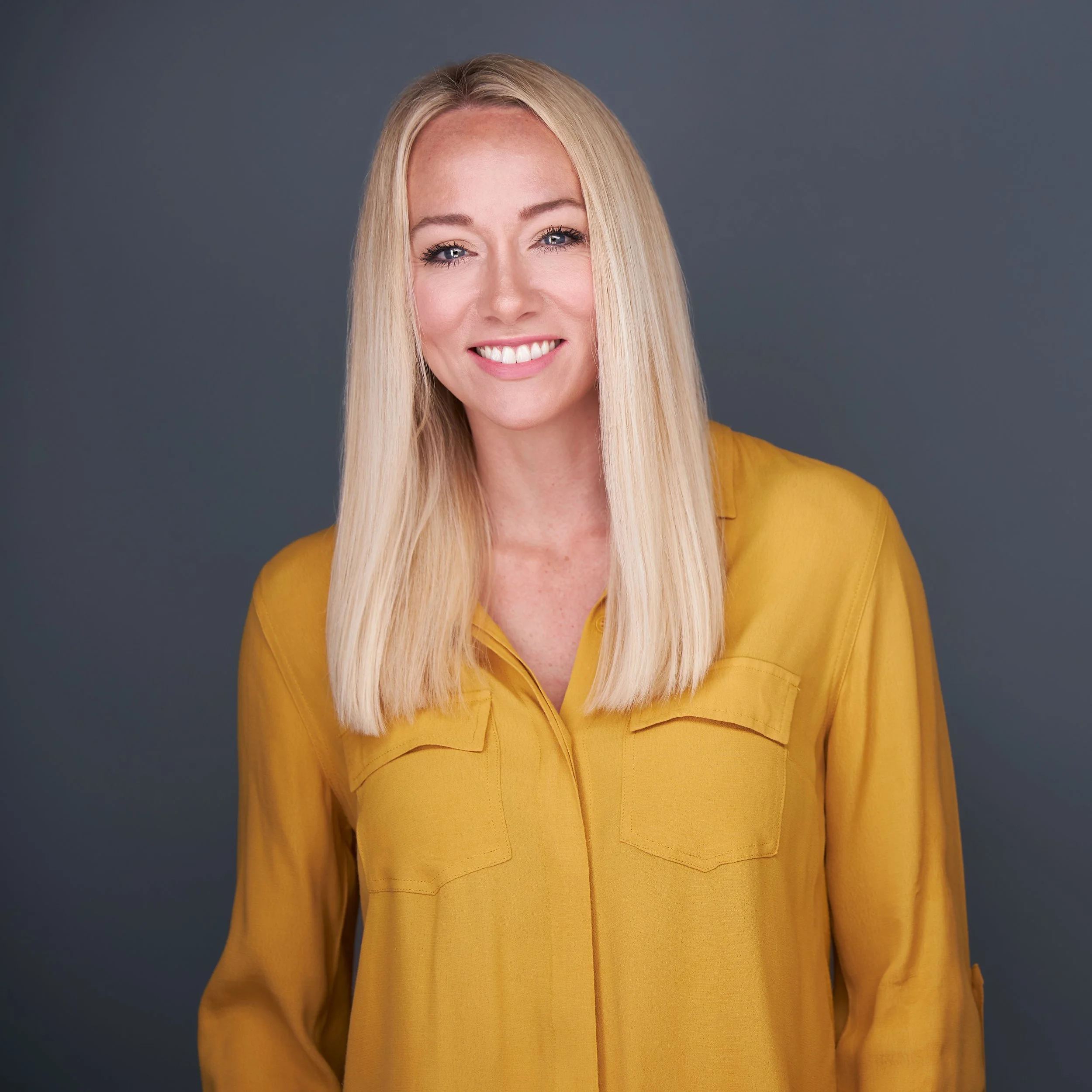 Close-up portrait of a woman in Scottsdale with long blonde hair, blue eyes, smiling, wearing a yellow top, against a gray background.