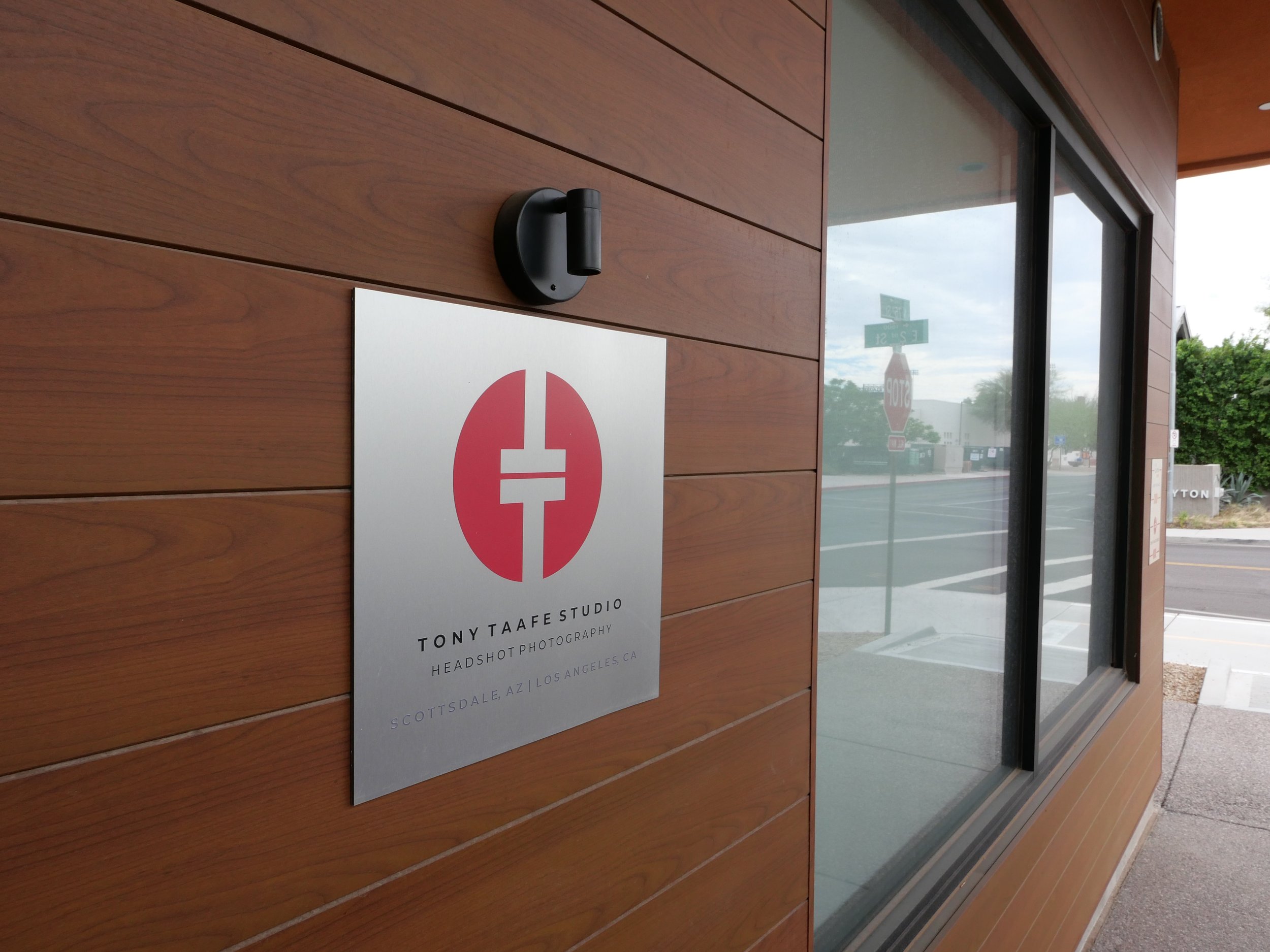 Wood-paneled exterior wall with a door, camera, and sign. The sign reads "TONY TAAFE STUDIO HEADSHOT PHOTOGRAPHY" with a location of Scottsdale, Arizona and Phoenix Arizona. The door has a large glass window showing a street and sidewalk outside.