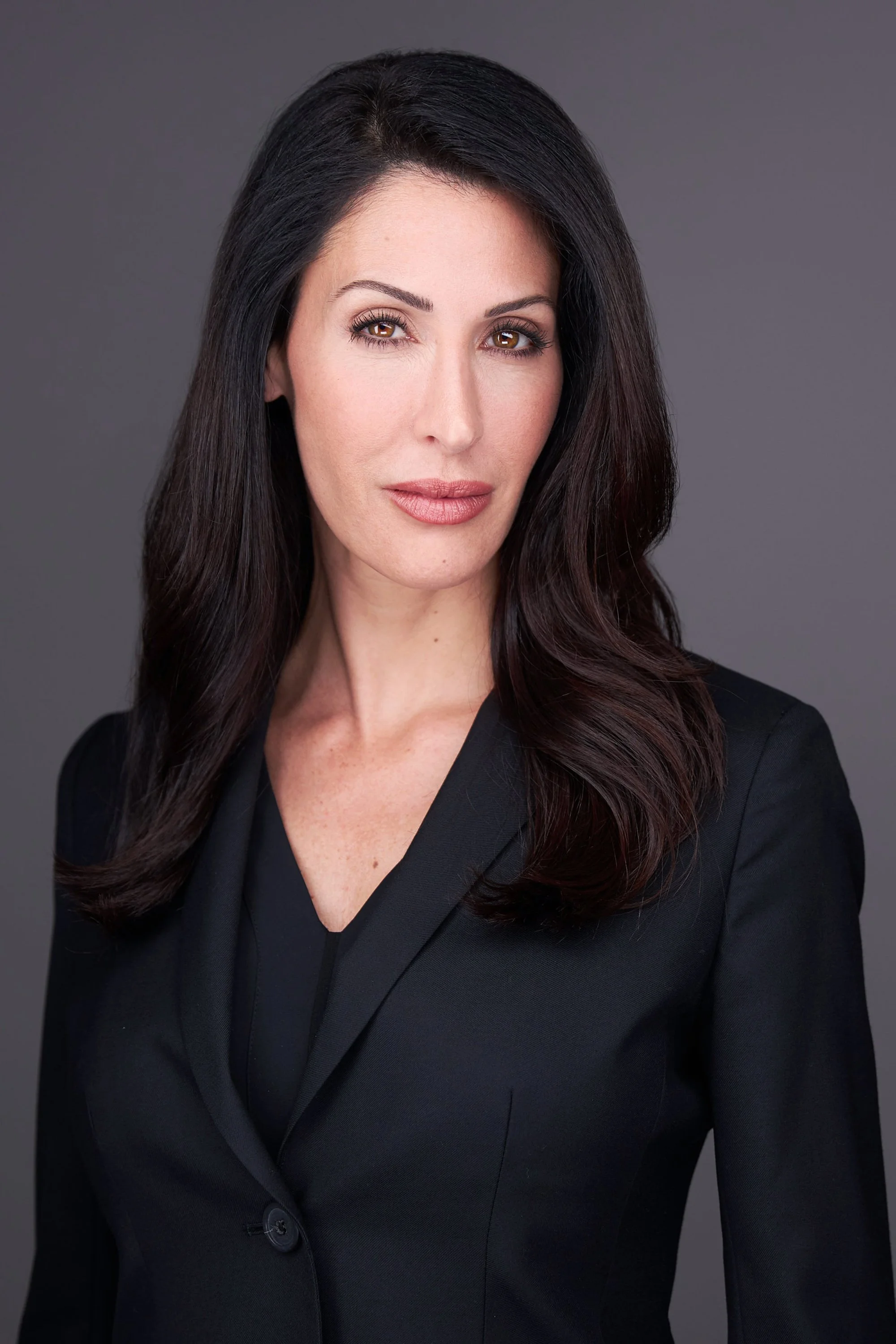 A confident woman with long dark hair, brown eyes, and a light lipstick, wearing a black blazer and shirt against a gray background.