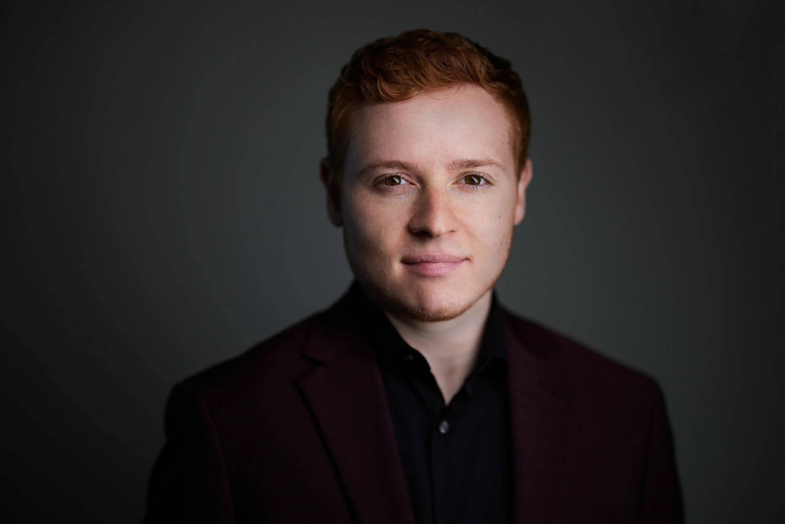 Portrait of a young man with short, curly red hair, wearing a dark blazer and black shirt, set against a dark gray background.