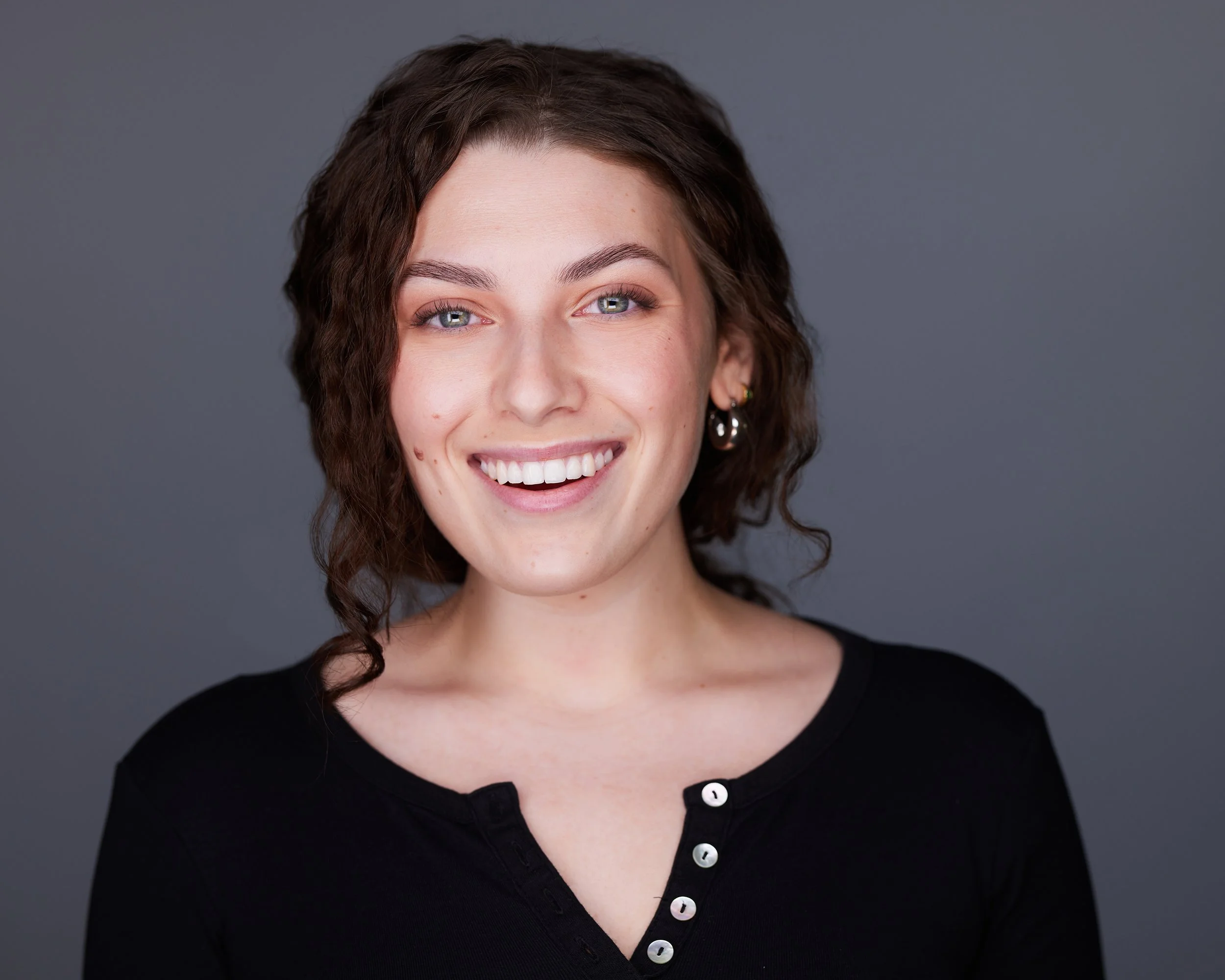 Headshot of a smiling woman with short, curly brown hair, blue eyes, and earrings, wearing a black button-up shirt against a gray background.