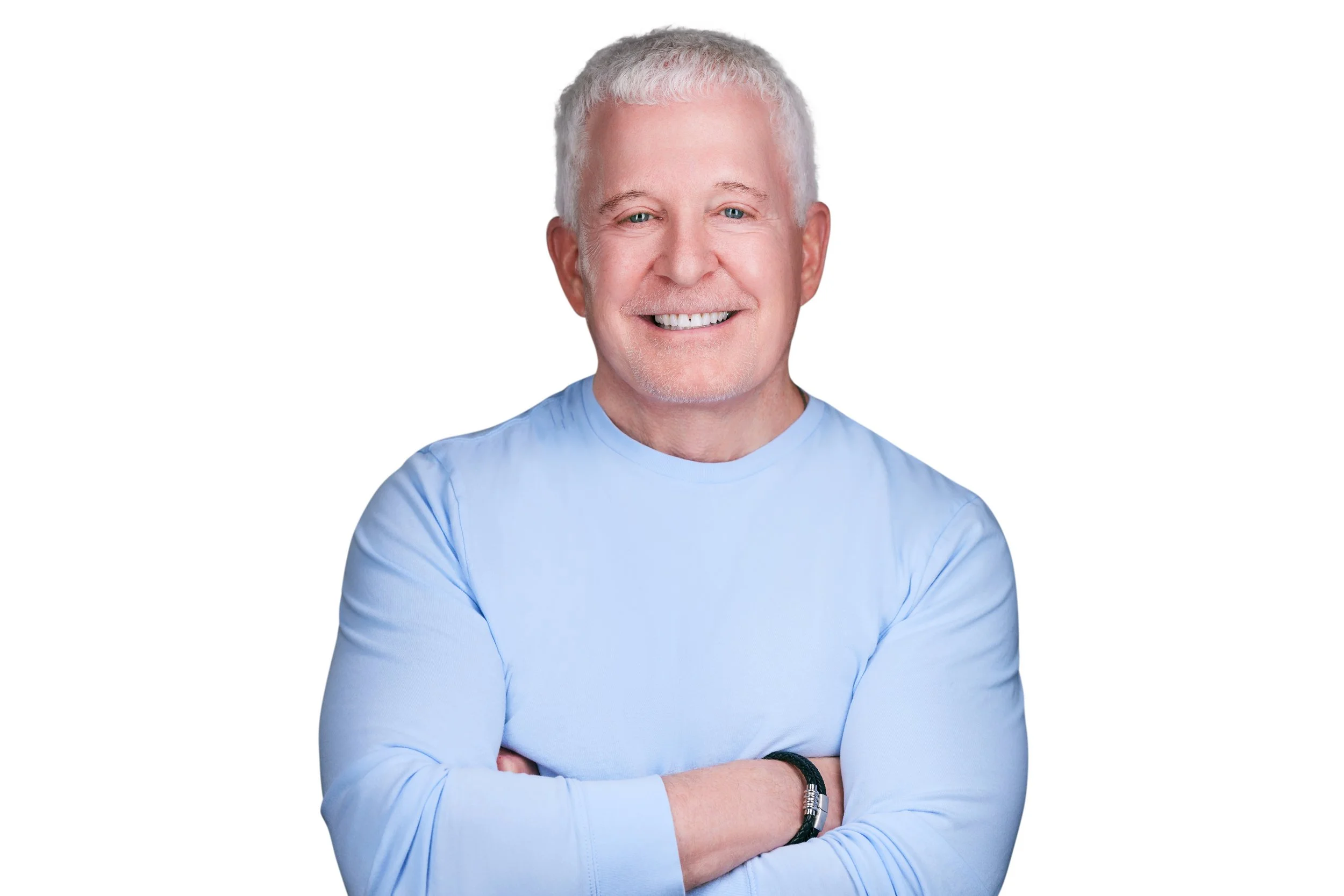 Smiling older man with gray hair and a beard, wearing a light blue shirt and a black bracelet, with arms crossed against a white background.
