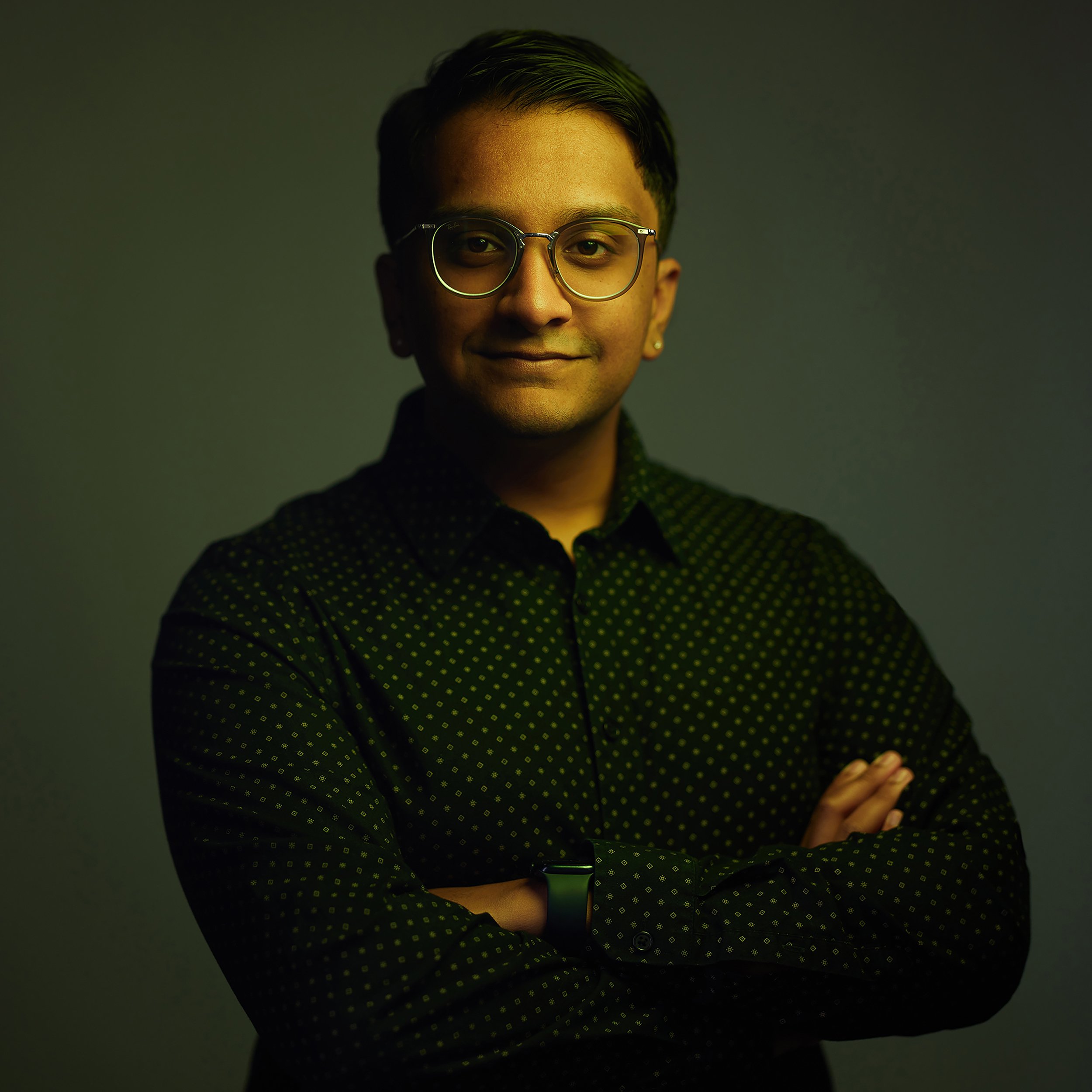 A technology entrepreneur having his headshot in Scottsdale by Tony Taafe, with short dark hair wearing round glasses, a black shirt with green polka dots, and a smartwatch, standing against a dark background with a subtle gradient.