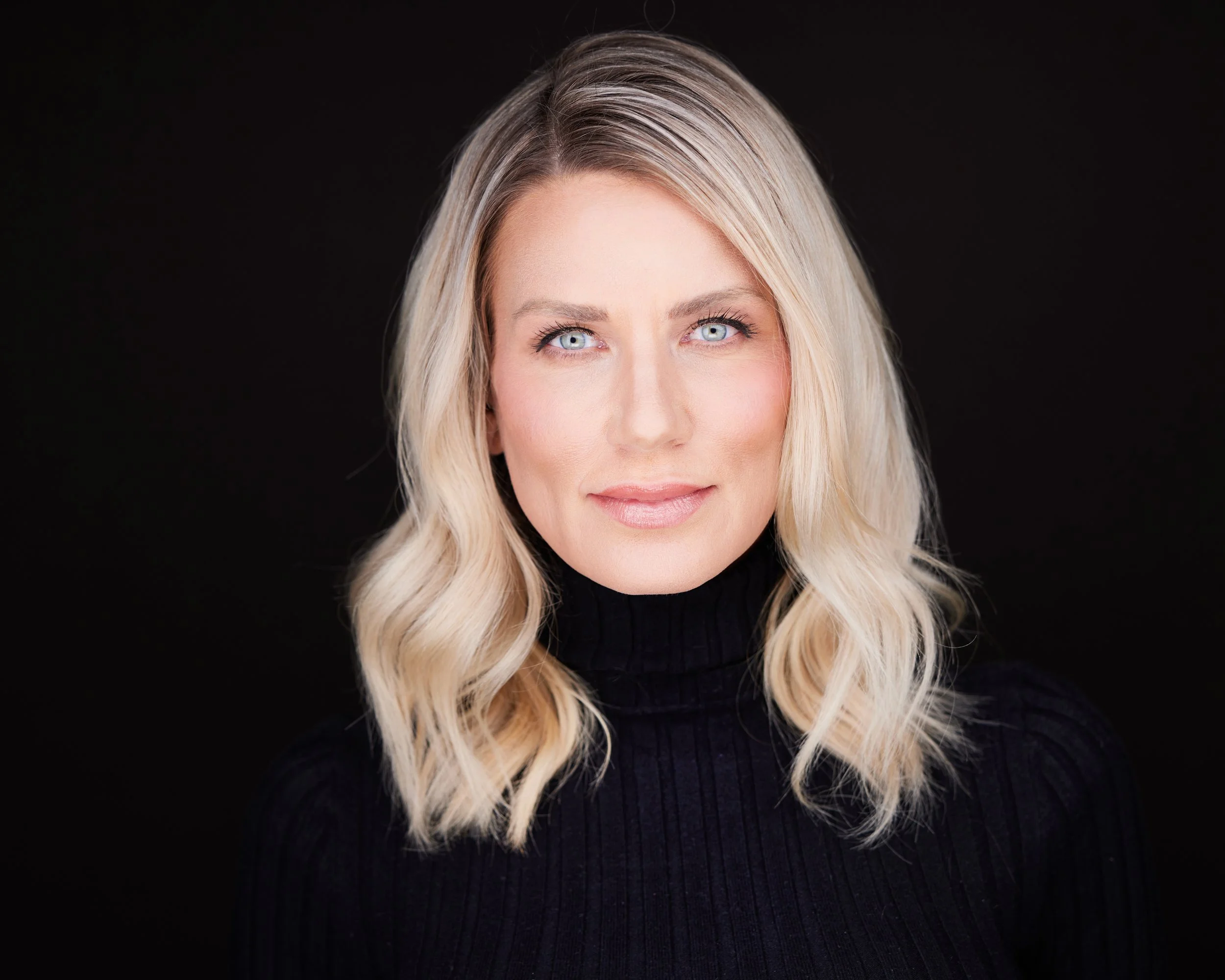 A woman with long, wavy blonde hair and blue eyes, wearing a black turtleneck top, against a black background.