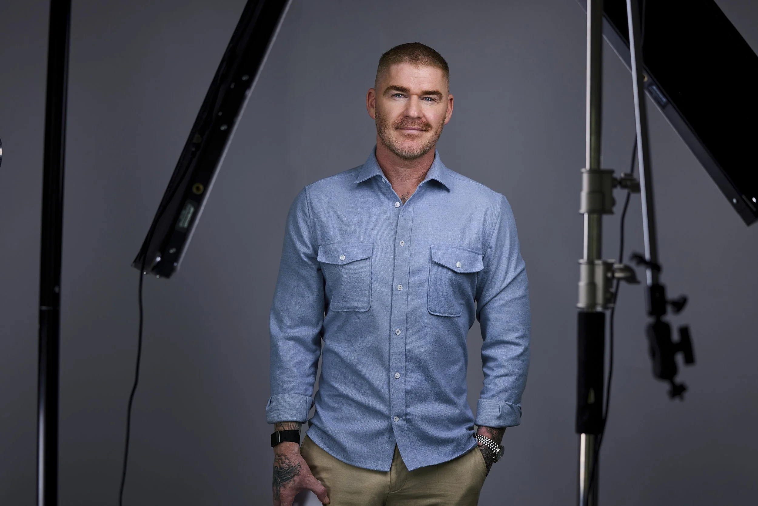 A man with short hair, a beard, and tattoos on his hands is standing in front of a gray background, wearing a light blue button-up shirt and beige pants, with his hands in his pockets. There are professional lighting and camera equipment visible around him.