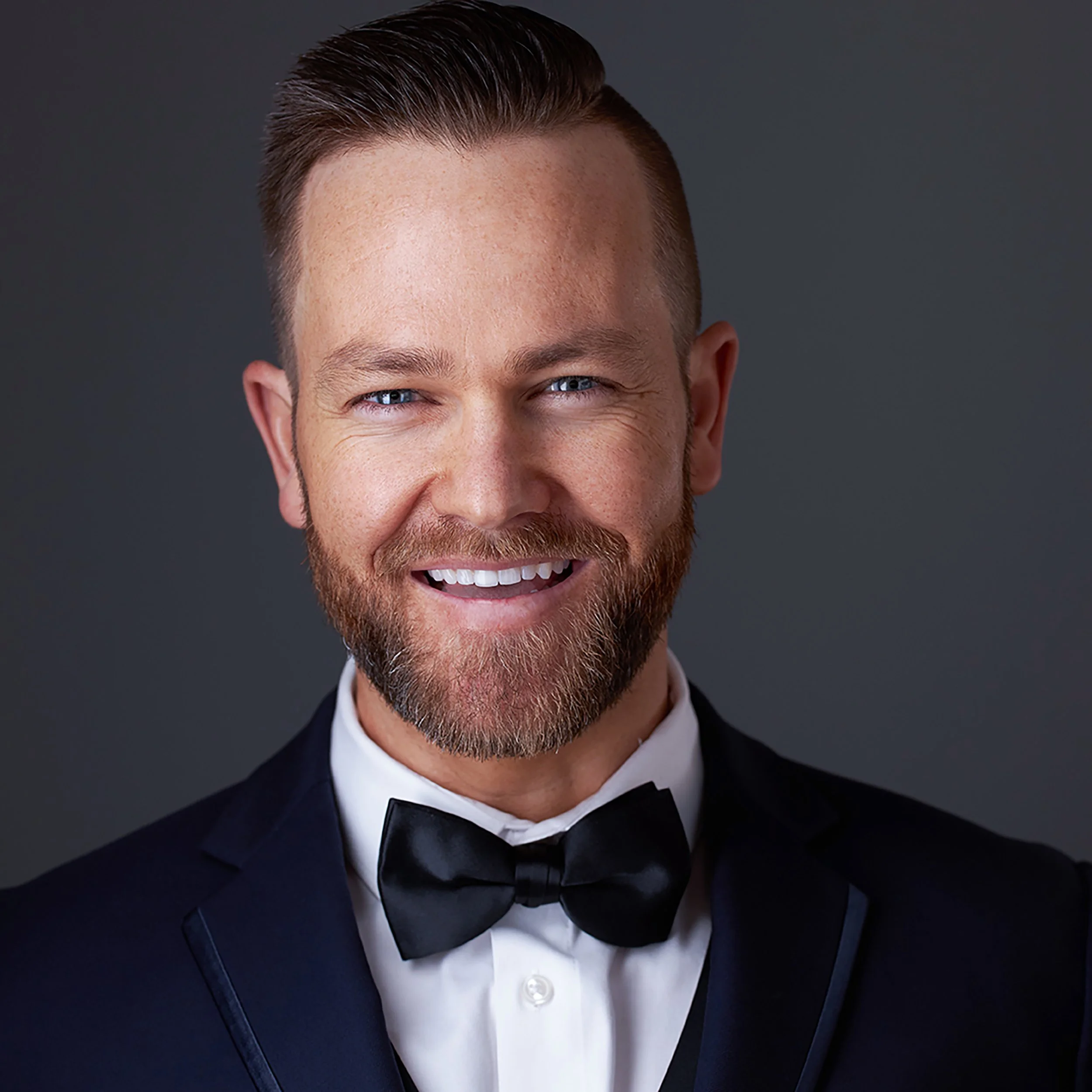 A close-up Headshot of a man taken in Scottsdale, with a neatly groomed hairstyle and beard, wearing a tuxedo with a white shirt and black bow tie, smiling against a dark gray background.