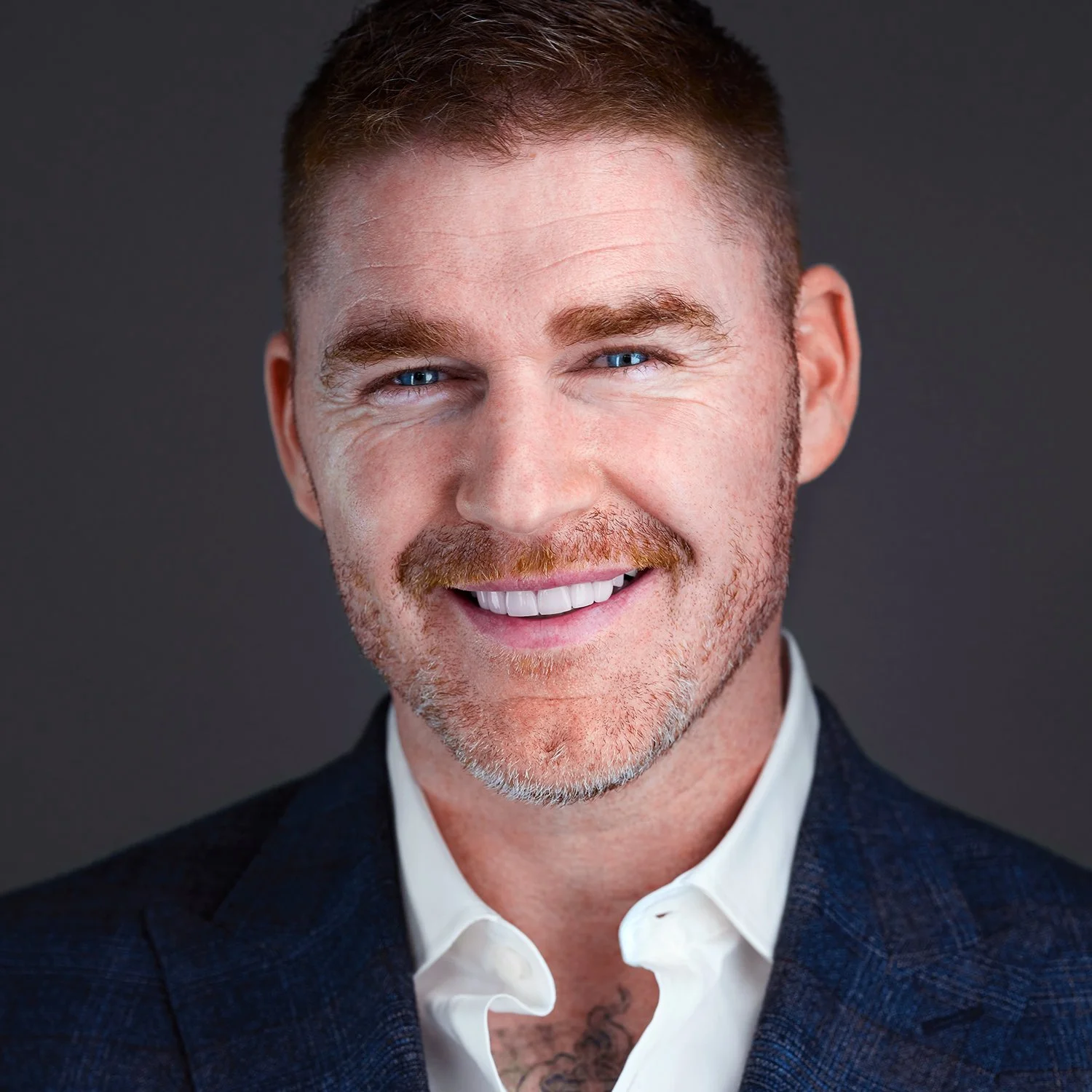 Close-up portrait of Tony Taafe with blue eyes, red hair, and a beard, wearing a dark blazer and white shirt against a dark background.