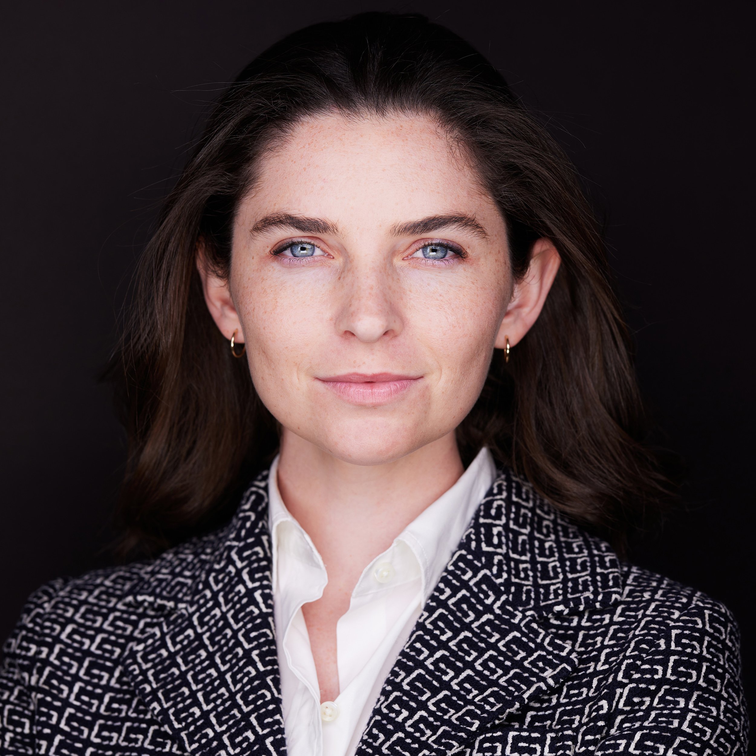 Close-up of a woman with blue eyes, dark hair, and light skin, wearing a white blouse and a black-and-white patterned blazer, against a black background.
