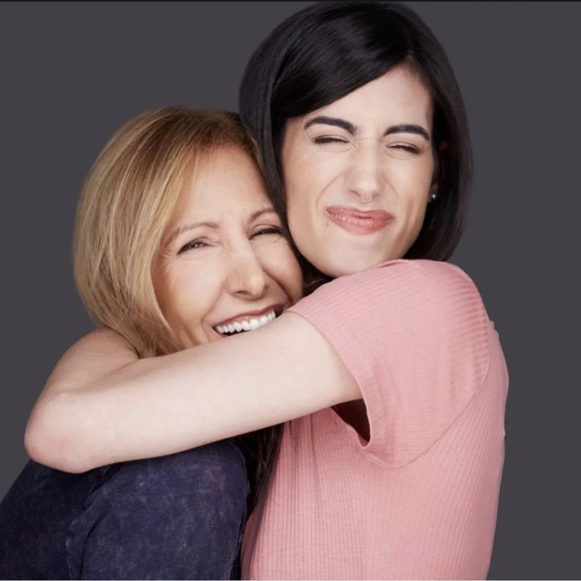 Two women hugging and smiling, one with black hair and another with blonde hair, against a dark grey background.