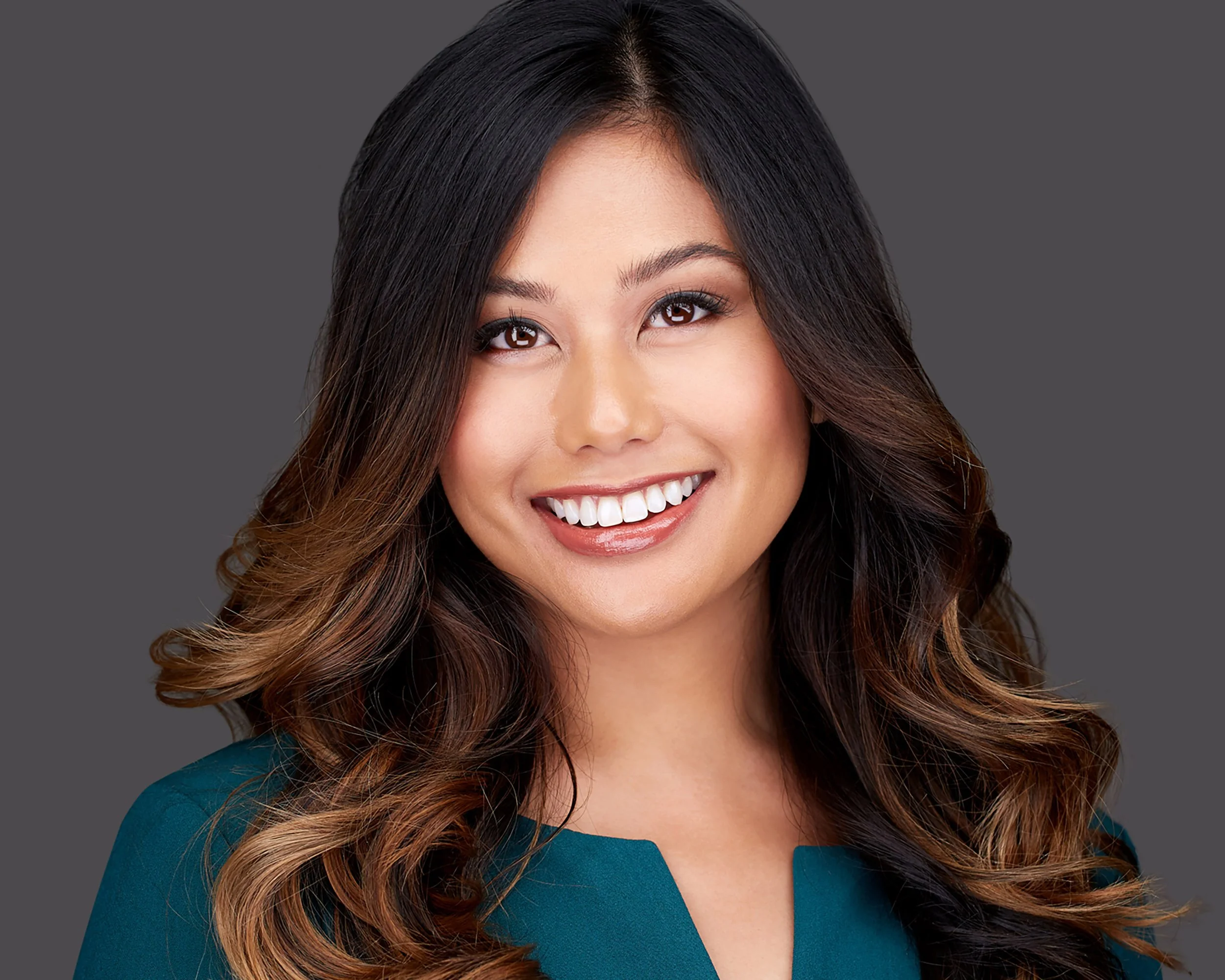 Headshot of a smiling woman with long wavy hair, wearing a teal top, against a gray background.