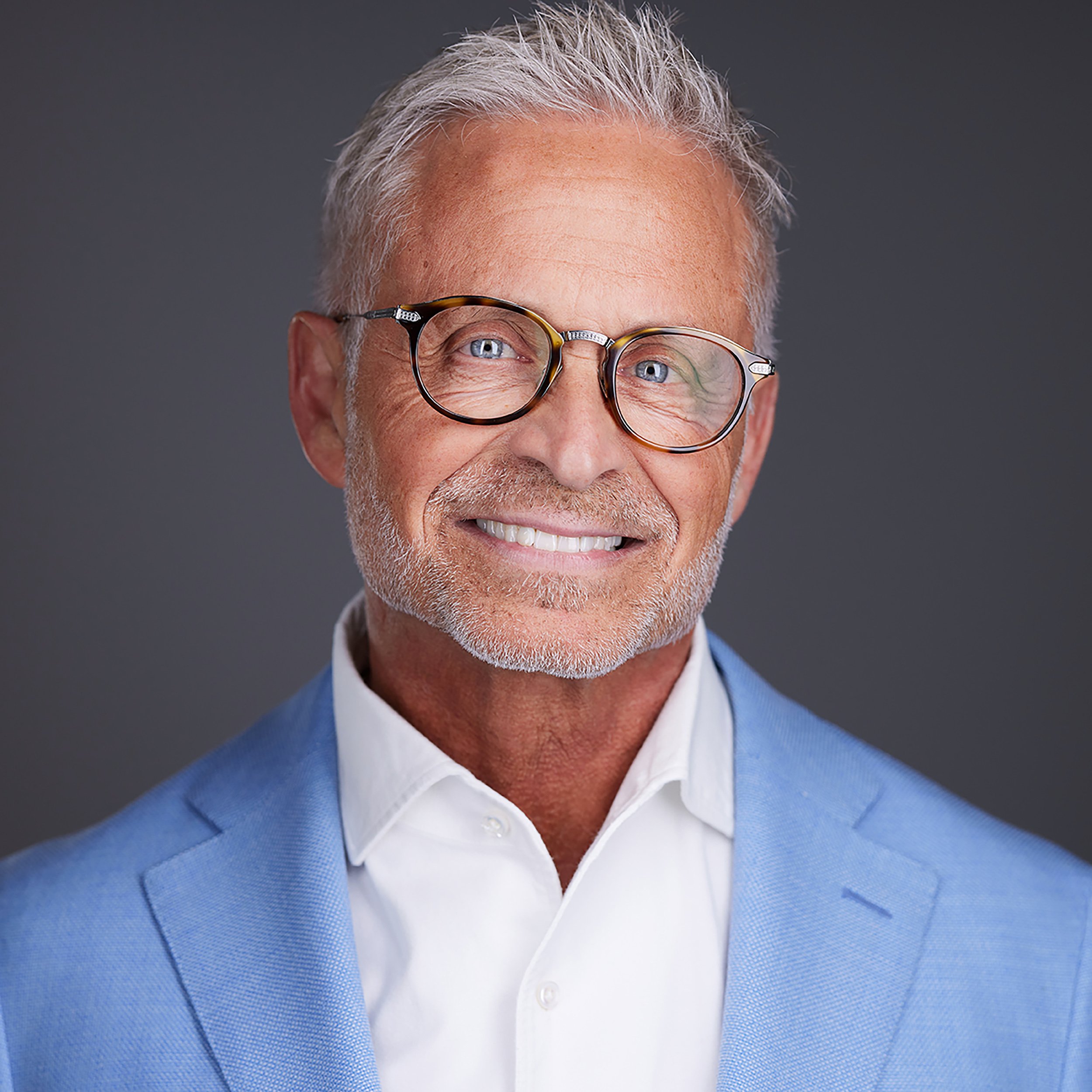 Headshot in Scottsdale of an older man with gray hair and a beard, wearing glasses, a white dress shirt, and a blue sweater, smiling against a dark background.