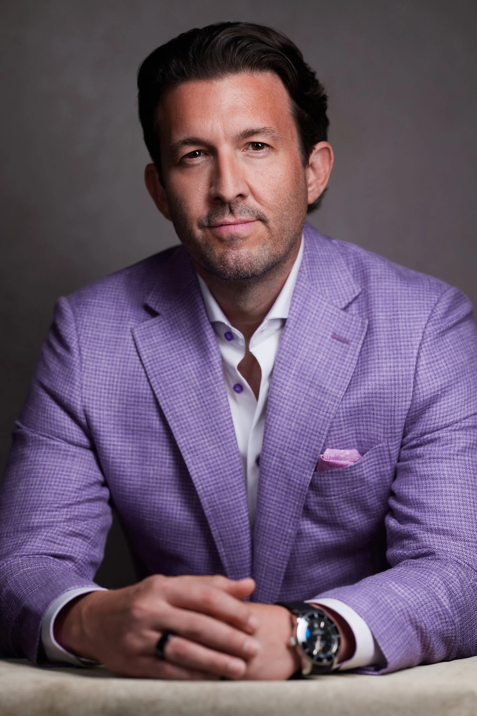 A man with dark hair and beard, wearing a purple checkered blazer over a white shirt, sitting at a table with his hands clasped. He is wearing a large watch on his wrist and a pink pocket square in his blazer pocket. The background is neutral gray.