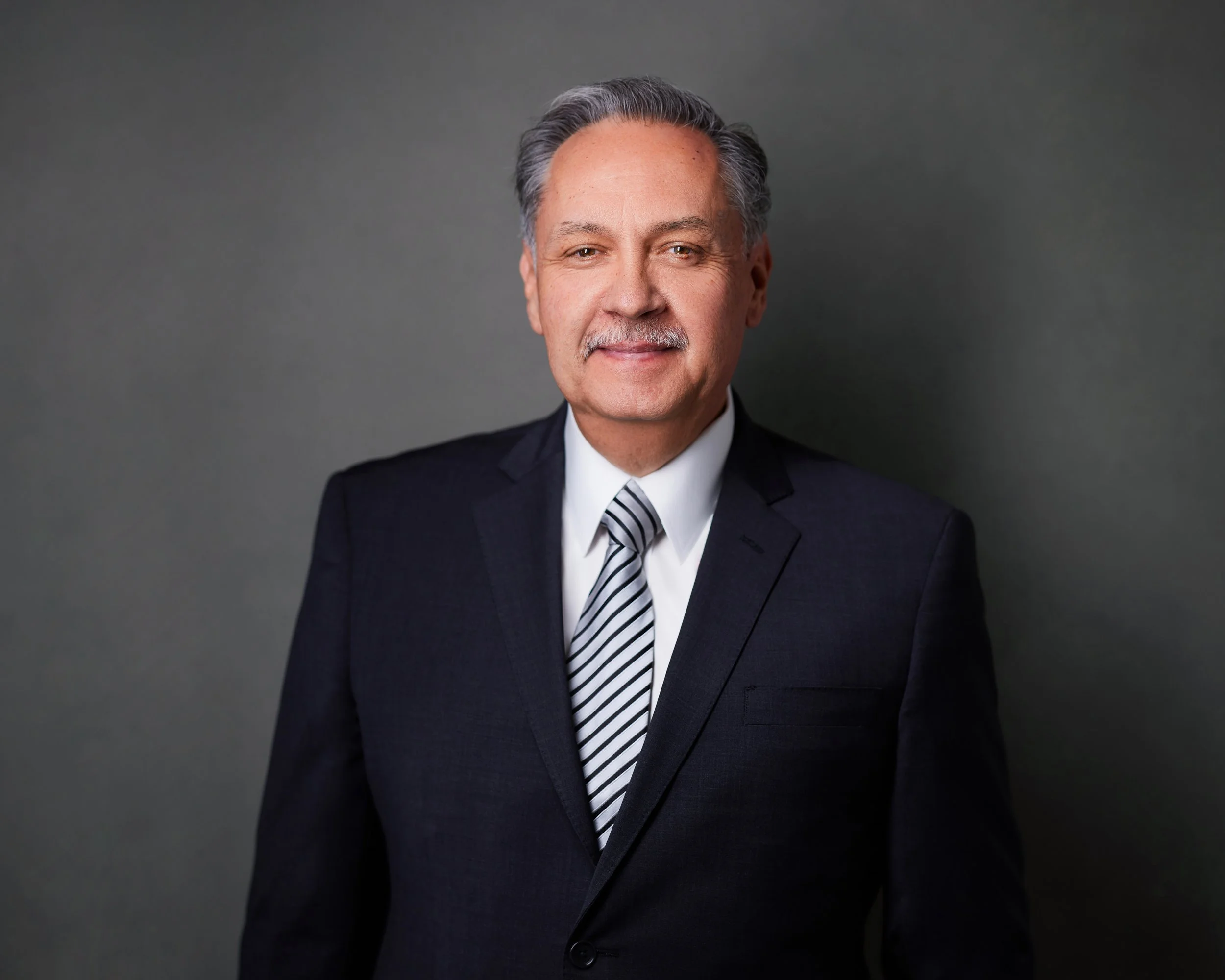 Headshot of a middle-aged man with gray hair, wearing a dark suit, white shirt, and striped tie, against a plain gray background.