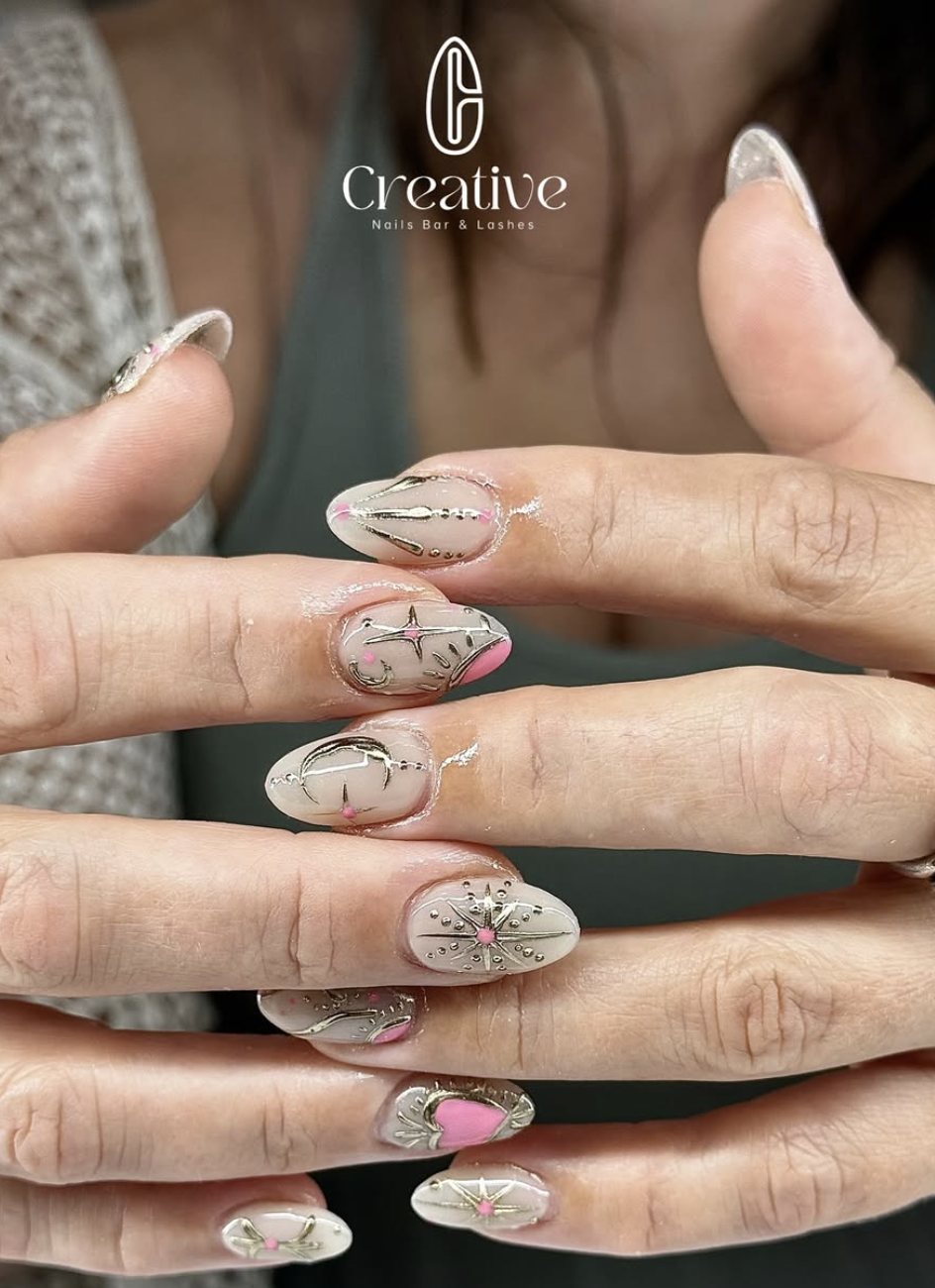 Close-up of a woman's hands displaying decorated nails with cream, pink, and gold nail art featuring small charms and designs, with a logo for 'Creative Nails Bar & Lashes' at the top.