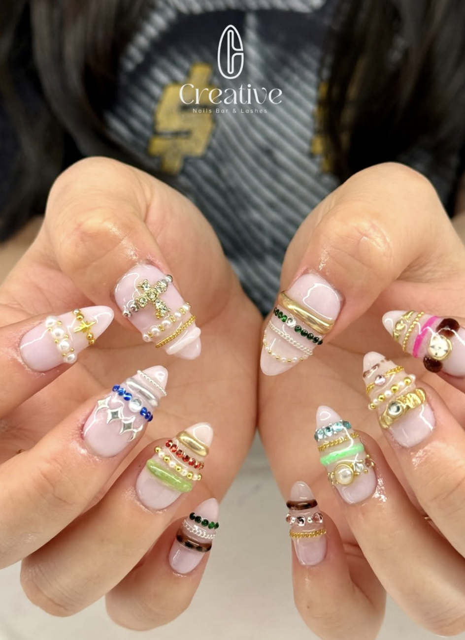 Decorative nail art with colorful rings, pearls, rhinestones, and tiny charms on long, oval-shaped nails, held by hands with a blurred background featuring a black apron with yellow patches and the word 'Creative'.
