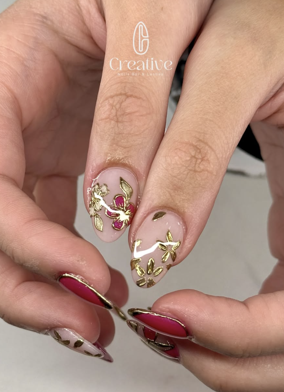 Close-up of a hand with manicured nails. The nails are decorated with pink and gold floral nail art and red polish with a gold edge.