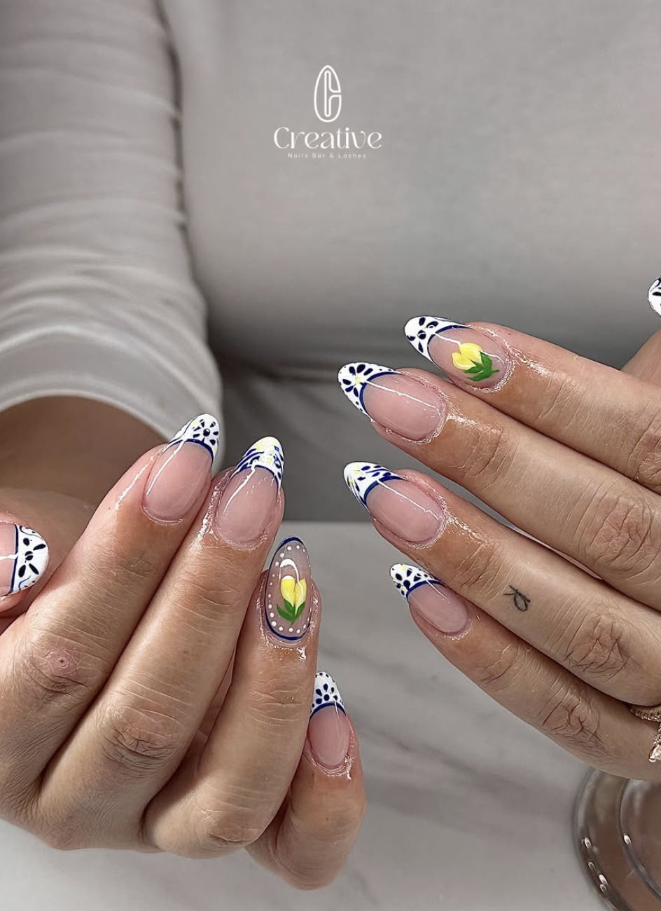 Close-up of hands with decorated nails featuring floral design, white tips with dark blue accents, and yellow flowers with green leaves, on a background with a logo that reads 'Creative Nails Bar & Lounge'.