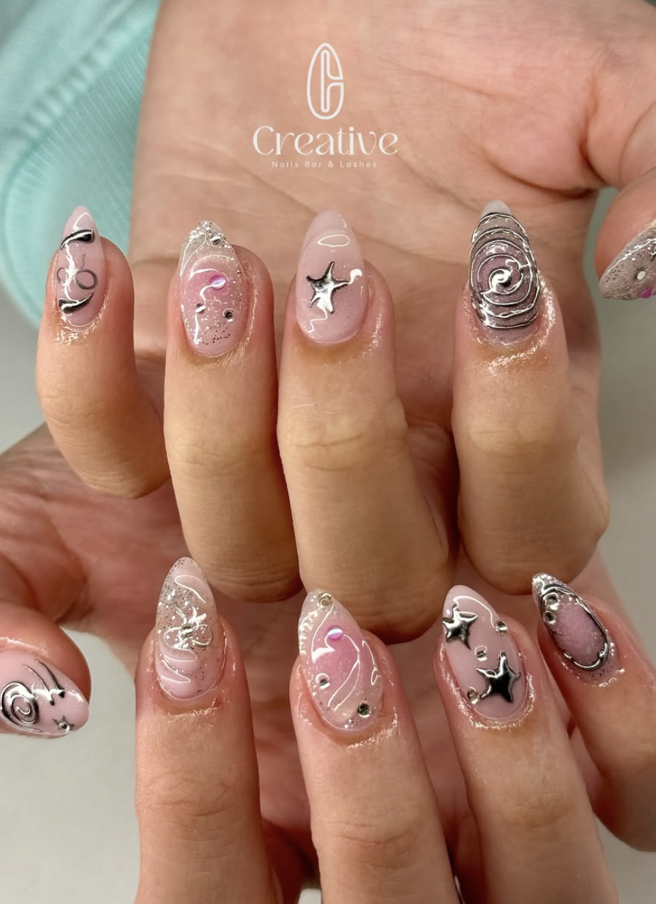 Close-up of a person's hand showing intricately designed nail art with silver star and moon embellishments, pink and silver swirled patterns, and glitter accents.