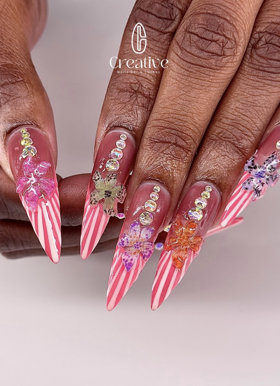 Close-up of a hand with long acrylic nails featuring pink and white striped designs, floral decals, and rhinestone embellishments.