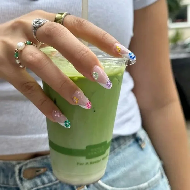 Person holding plastic cup filled with green beverage, standing outdoors, wearing colorful nail art and multiple rings.