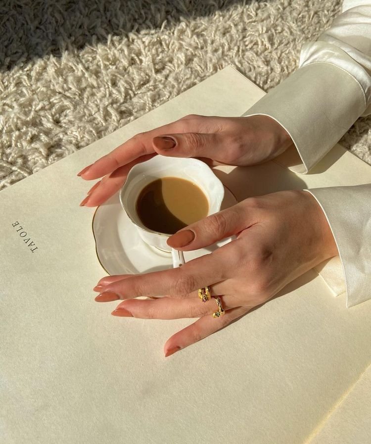 Two hands with manicured nails, one with gold jewelry, holding a small white cup of coffee or espresso over a white table with a cream-colored textured carpet in the background.