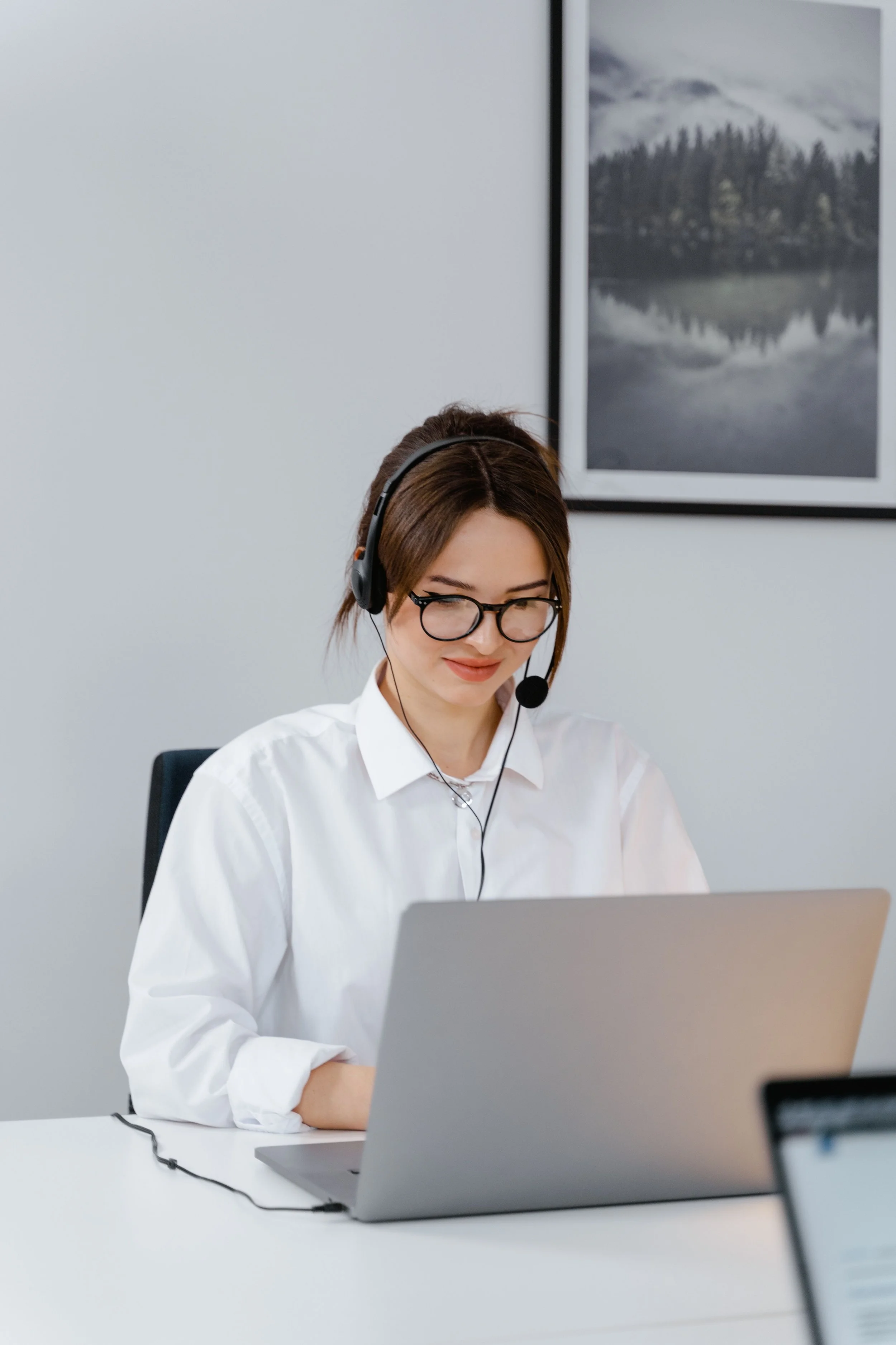A woman with brown hair, glasses, and a headset smiling while working at a laptop in an office.