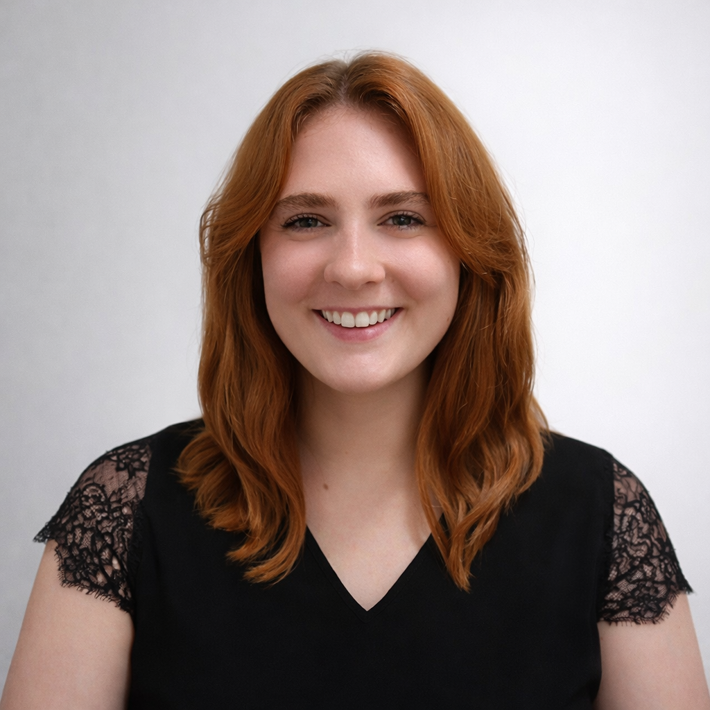 Portrait of a young woman with red hair wearing a black top with lace sleeves, smiling against a light plain background.