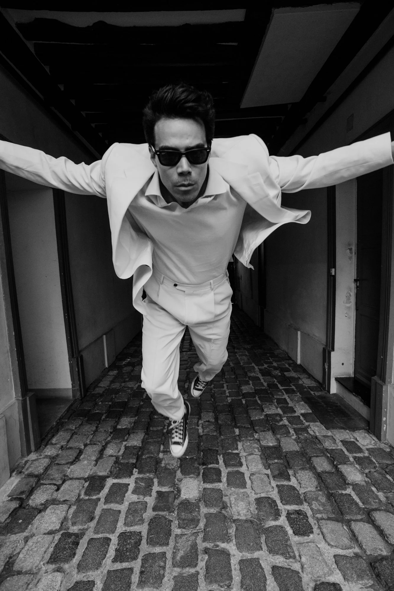 A man in a white suit, sneakers, sunglasses, and a collared shirt crawling or walking through a doorway, with arms outstretched and intense expression.