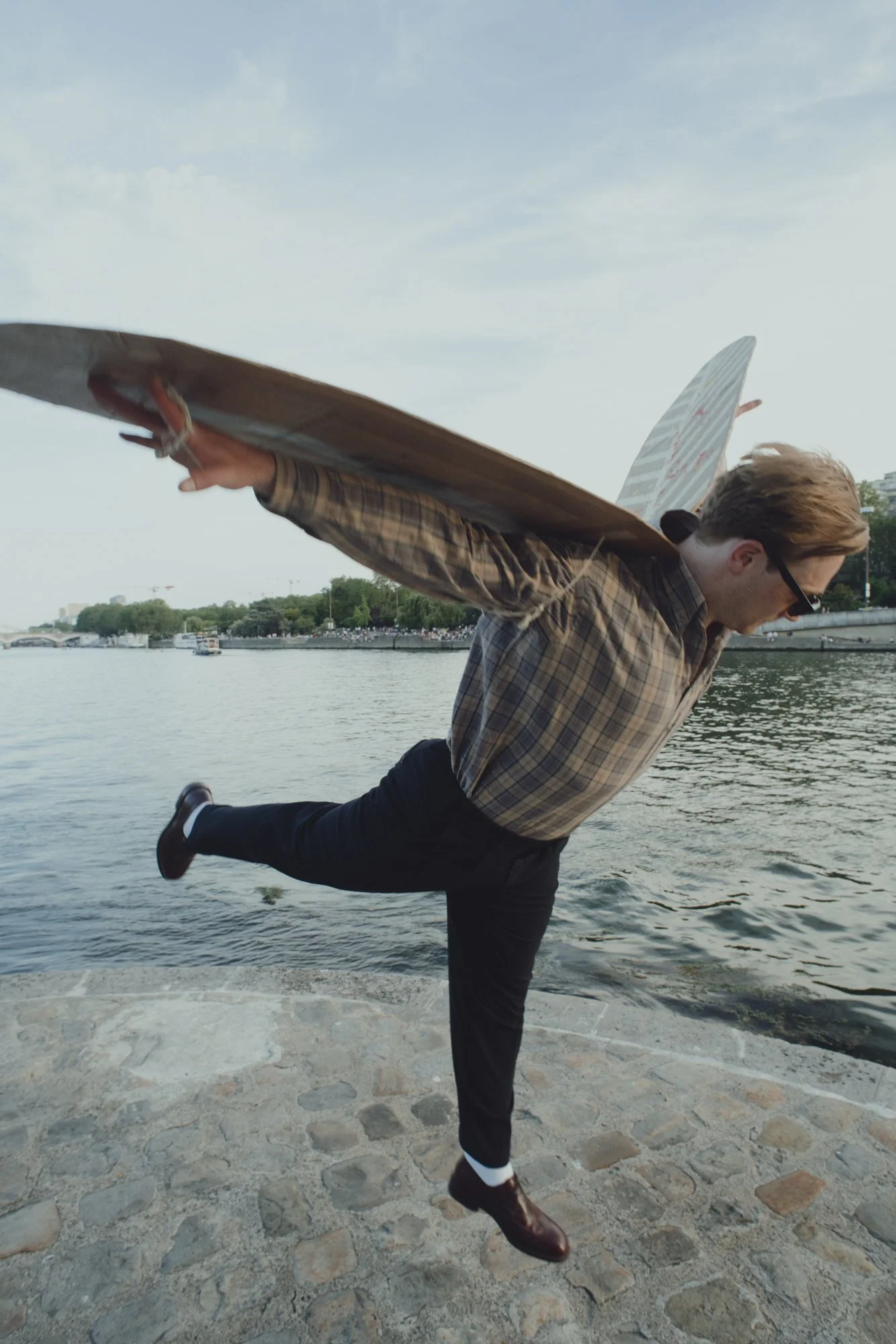 A man with glasses and a plaid shirt is holding a skateboard on his back while balancing on one foot on a stone-paved area near water. The man is dressed in black pants and brown boots, and appears to be doing a dance move or balancing pose near a river, with trees and city buildings in the background.