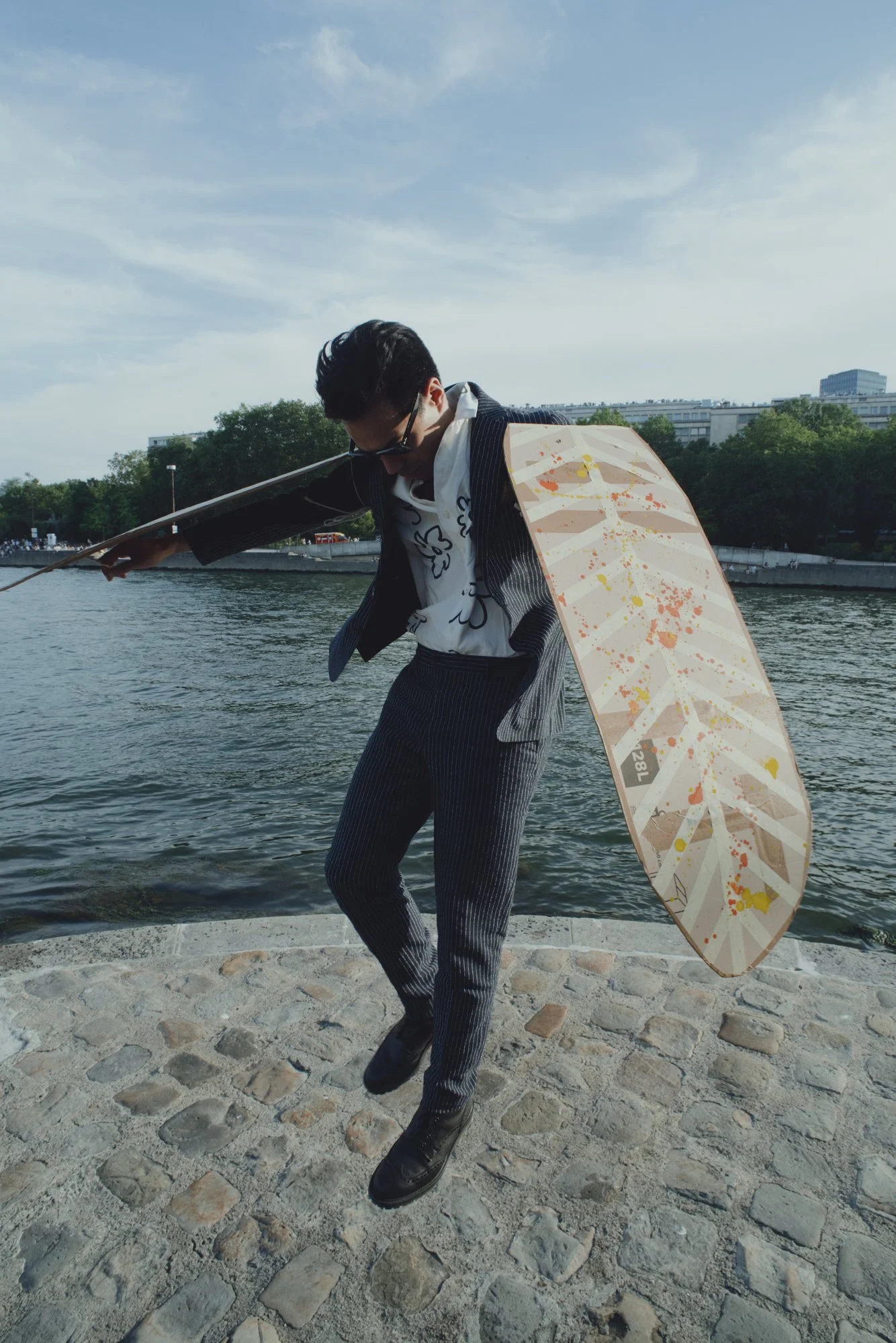 A man dressed in a black blazer, white shirt with black illustrations, and striped pants is holding a large skateboard with a beige and splattered paint design, standing on a cobblestone area by the water with trees and buildings in the background.