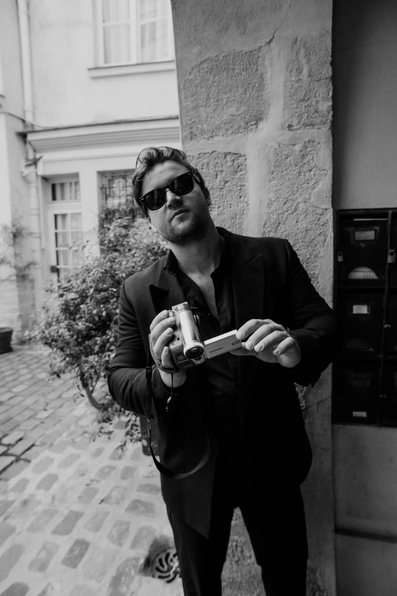 A man wearing sunglasses and a suit, holding a camera, standing outside near a stone wall with buildings and bushes in the background.
