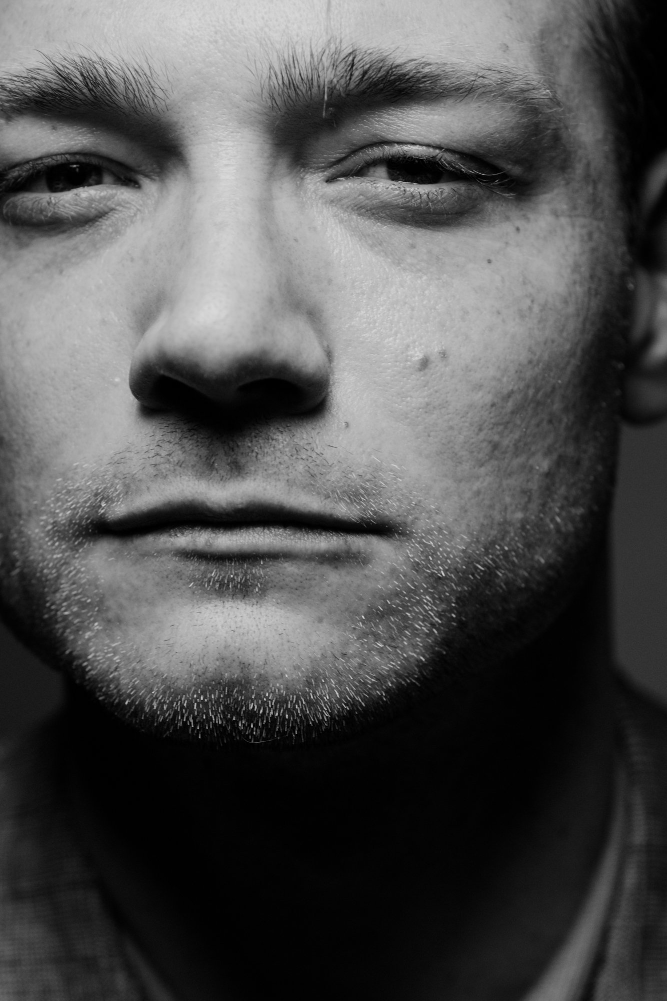 Close-up black and white photo of a person's face, with one eye closed and the other eye open, featuring detailed skin texture, eyebrows, and facial hair.