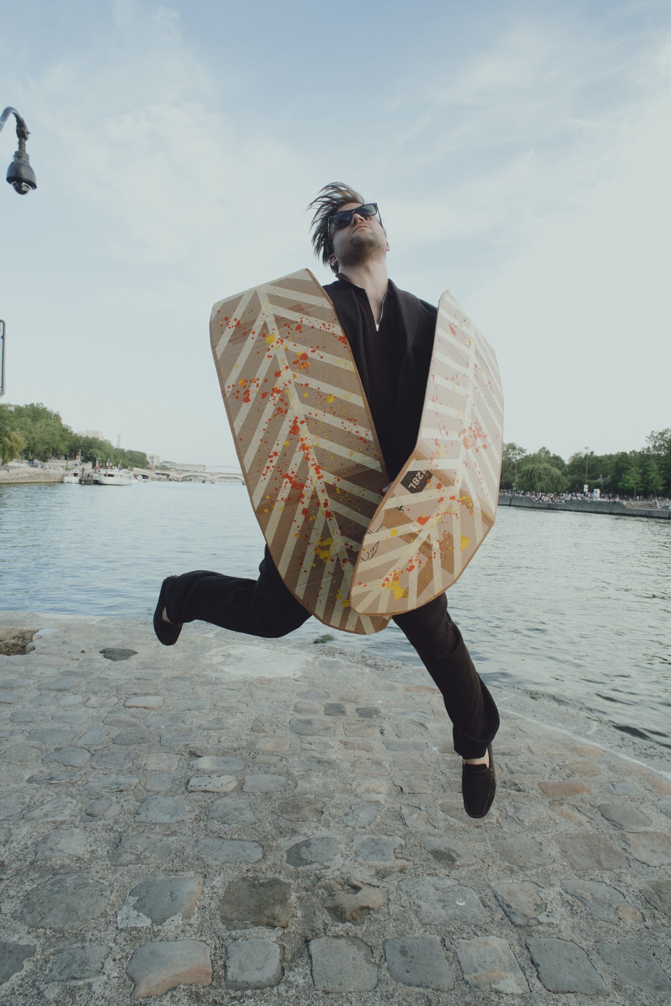 A man wearing sunglasses and a black outfit is jumping in the air with one leg bent and the other extended, near a river with boats and greenery in the background. He is holding an oversized cardboard object with a colorful, abstract design. The sky is mostly clear with some clouds.