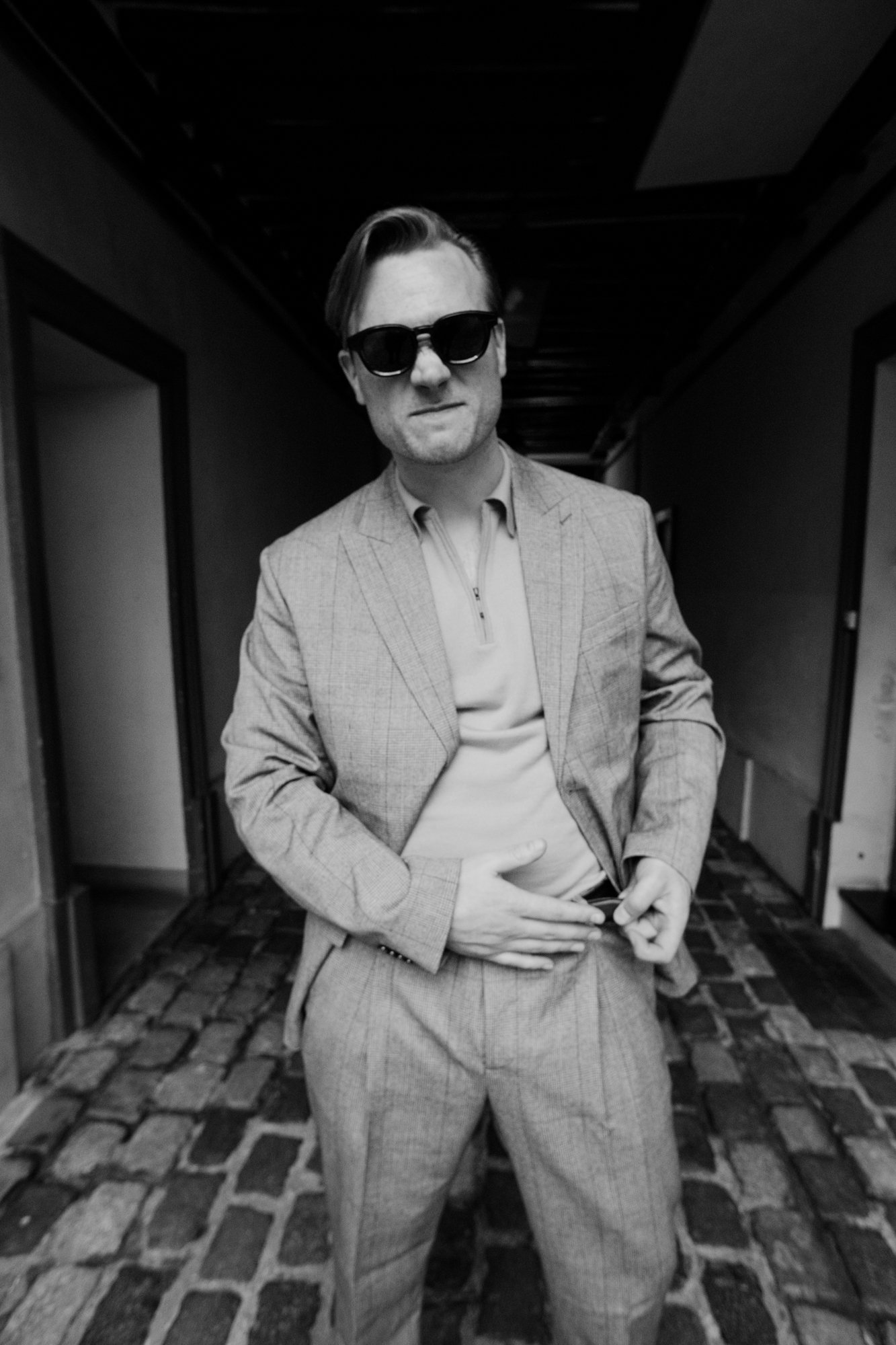 A man wearing sunglasses and a light-colored suit standing in a corridor with dark lighting and cobblestone floor.