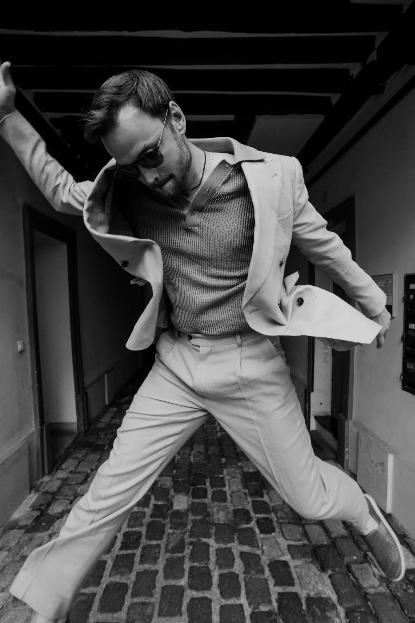 A man in a suit and sunglasses striking a dramatic pose in a hallway, with one arm raised and one leg stretched out.