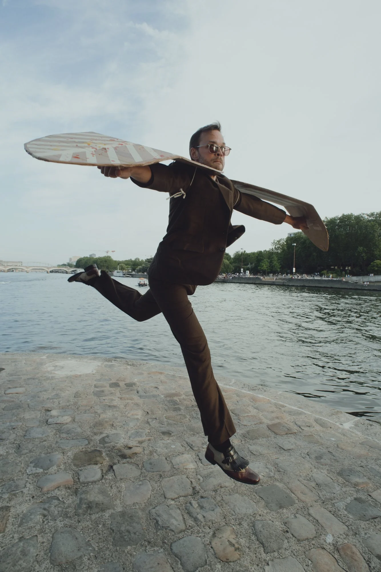 A man dressed in business attire is running near a riverbank with a large cardboard surfboard on his back. He appears to be in motion, with one leg lifted and bent at the knee, and the other supporting his stance. The background includes a river, some boats, a bridge, and trees along the shoreline under a partly cloudy sky.